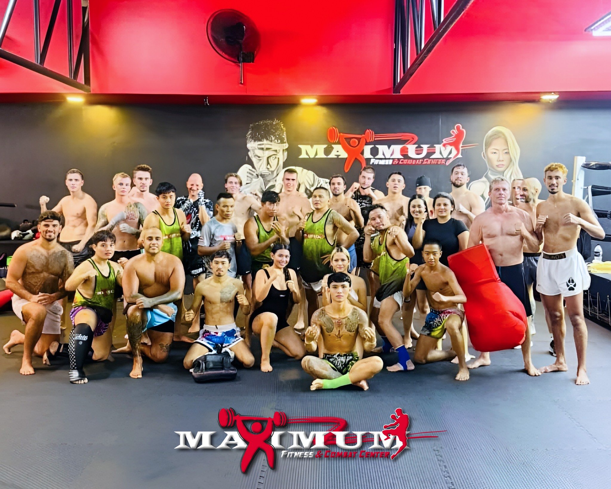 Group of mixed martial arts fighters posing together in a fitness center with a large logo on the back wall that reads 'Max Muay' and features a graphic of a person lifting weights and a female martial artist. Many are in fighting shorts or training 