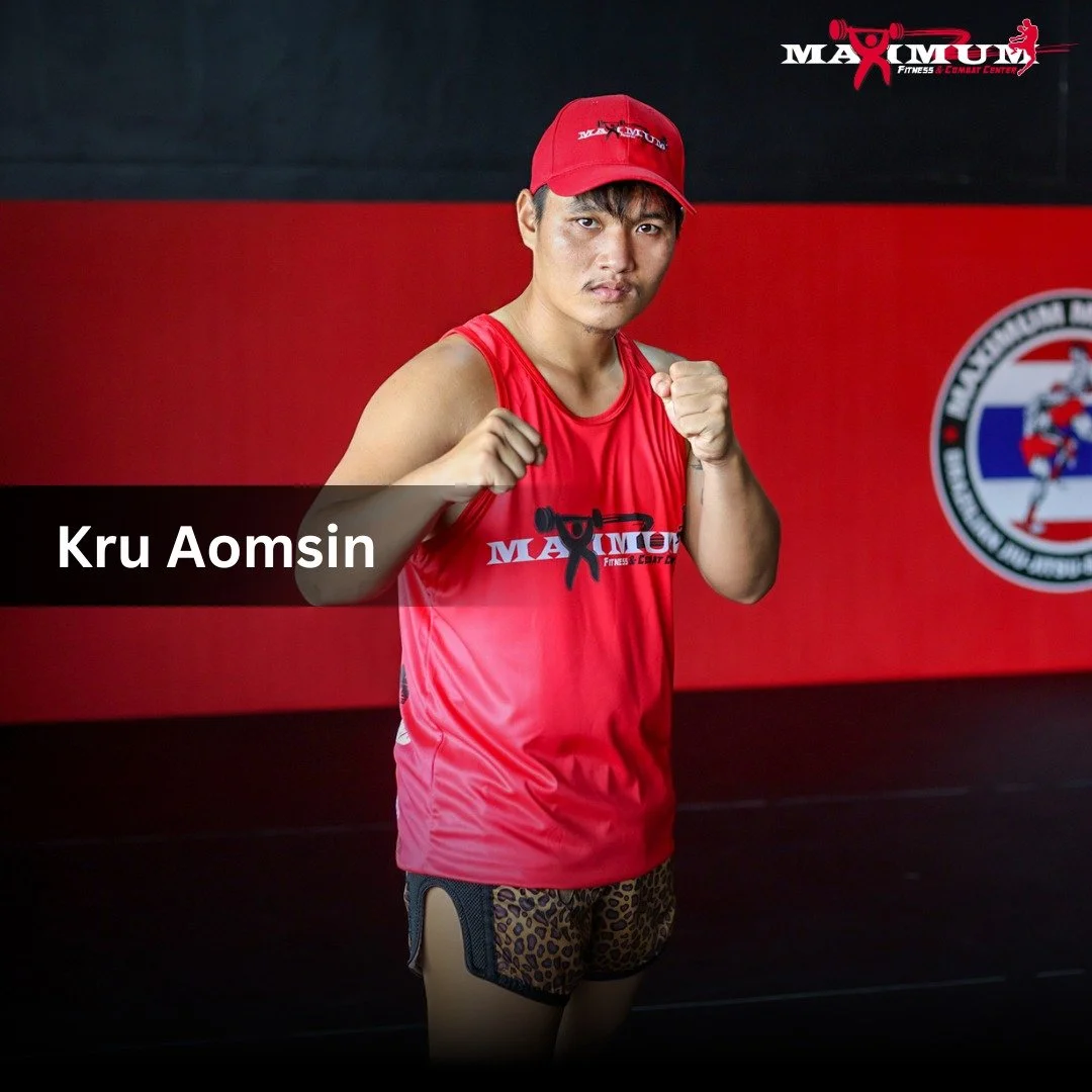 A male Muay Thai fighter standing in a gym holding his fists up in a fighting stance. He wears a red tank top and matching cap, leopard print shorts, and is in front of a red and black wall with a fitness center logo.