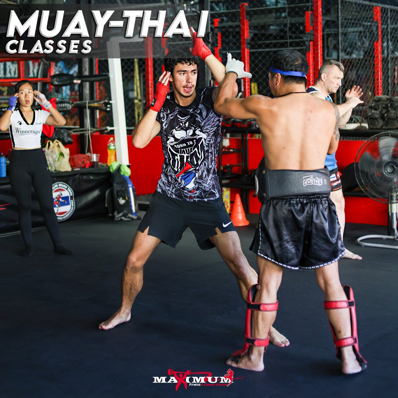 Two men sparring in Muay Thai training, with one in black shorts and black protective gear and the other in black shorts with red hand wraps, in a gym with red accents and other people in the background.