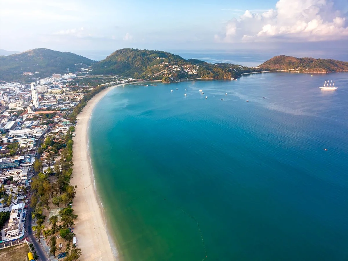 Aerial view of a crescent beach along a city coastline with buildings, hills in the background, and boats in the water.