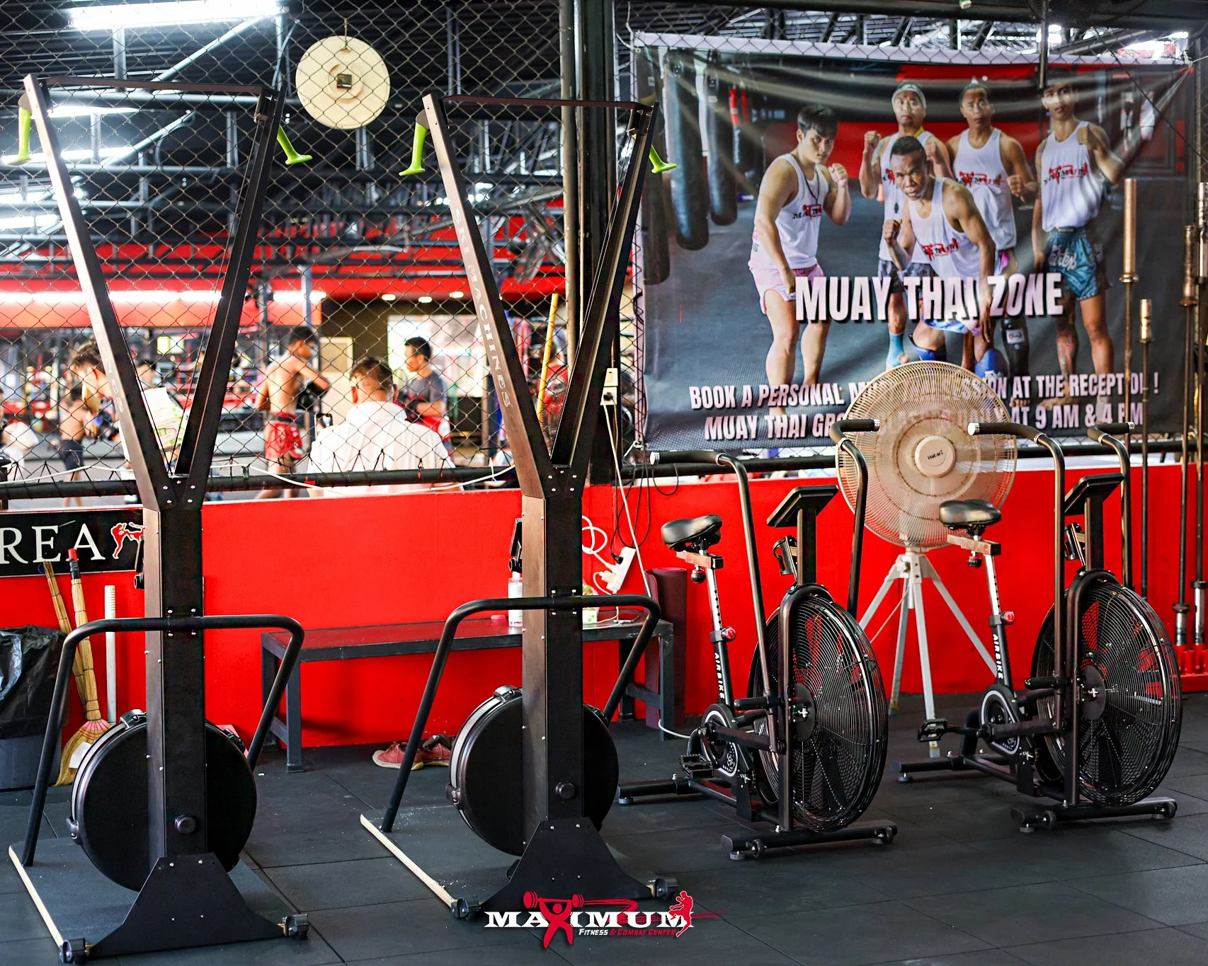 Gym area with fitness equipment, including two air bikes and a fan, in front of a boxing gym with people training in the background. A large poster advertises Muay Thai classes.