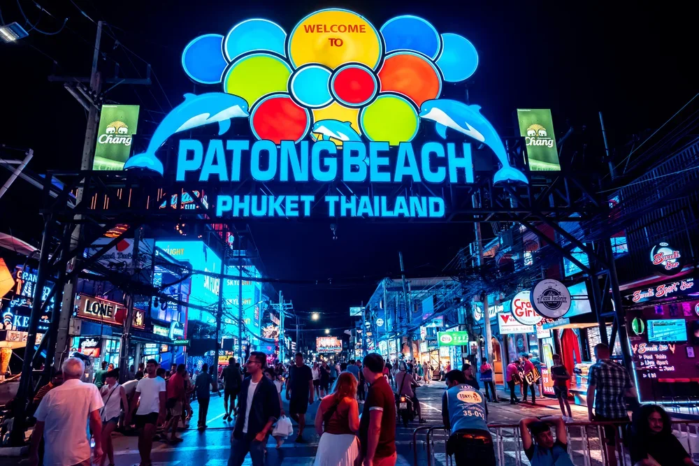 Nighttime scene of Patong Beach in Phuket, Thailand with neon signs, a large illuminated sign reading "Welcome to Patong Beach Phuket Thailand," and crowds of people walking along the lively street.