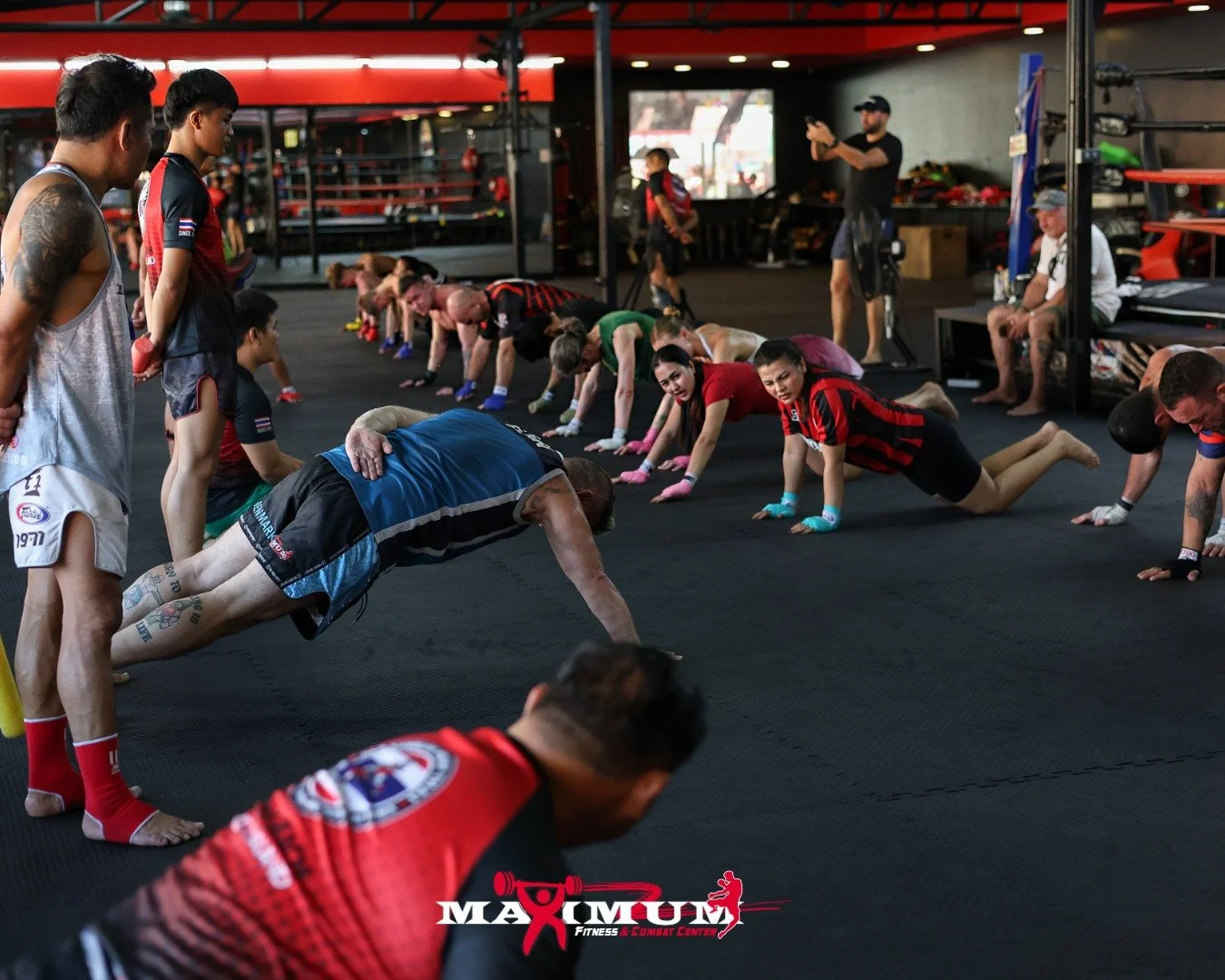 A group of people doing push-ups on a black gym floor during a fitness class at Maxmum Fitness & Combat Center, with some instructors observing and guiding.