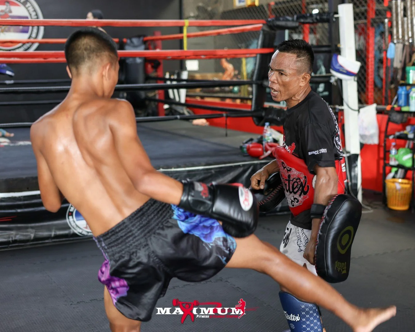 A shirtless male martial artist practicing kickboxing with a trainer holding focus pads in a gym with a boxing ring in the background.