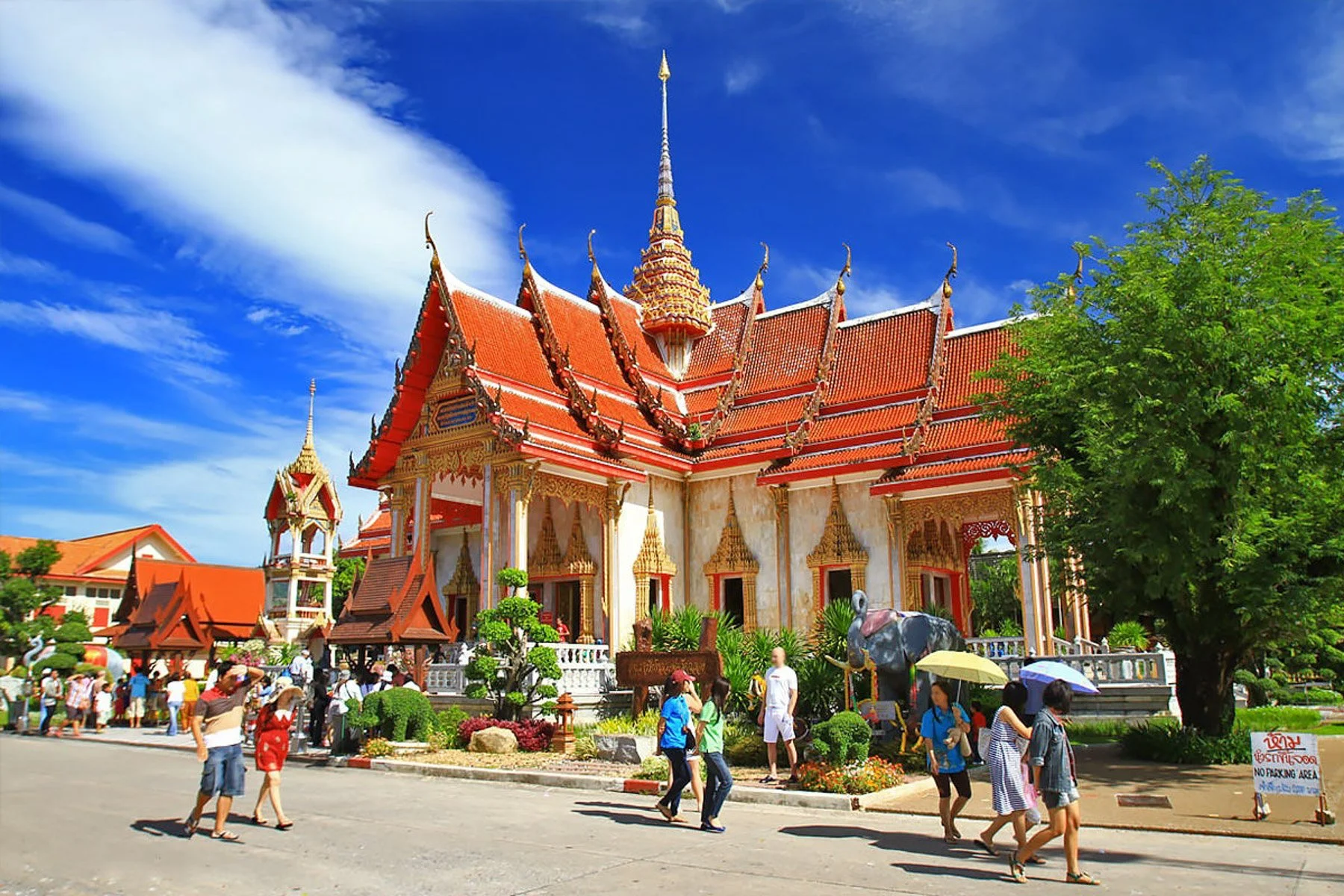Traditional Thai temple with ornate architecture, red and gold roof, and a tall spire, surrounded by visitors and lush greenery under a bright blue sky.