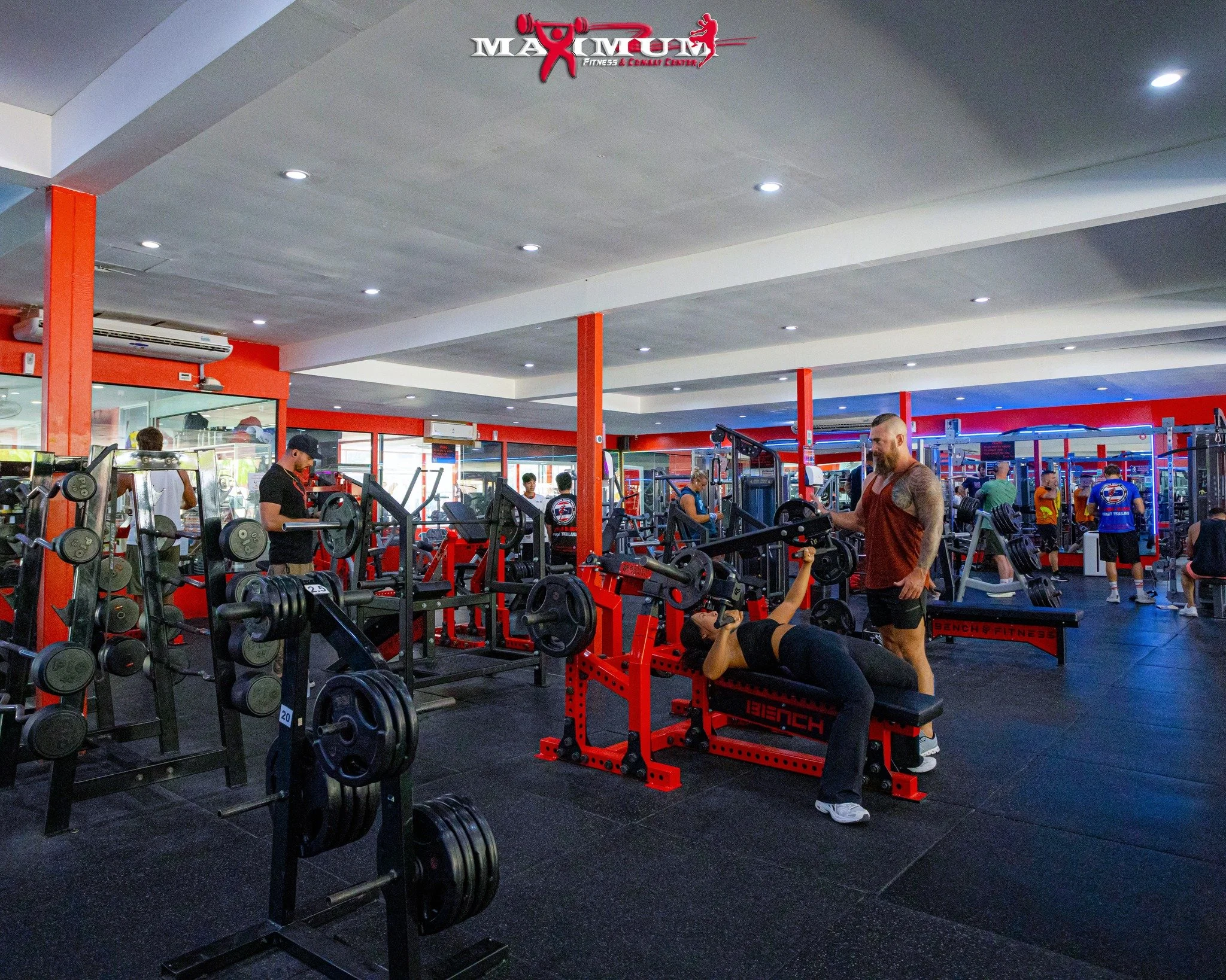People working out with weights and fitness equipment inside a gym with red accents.