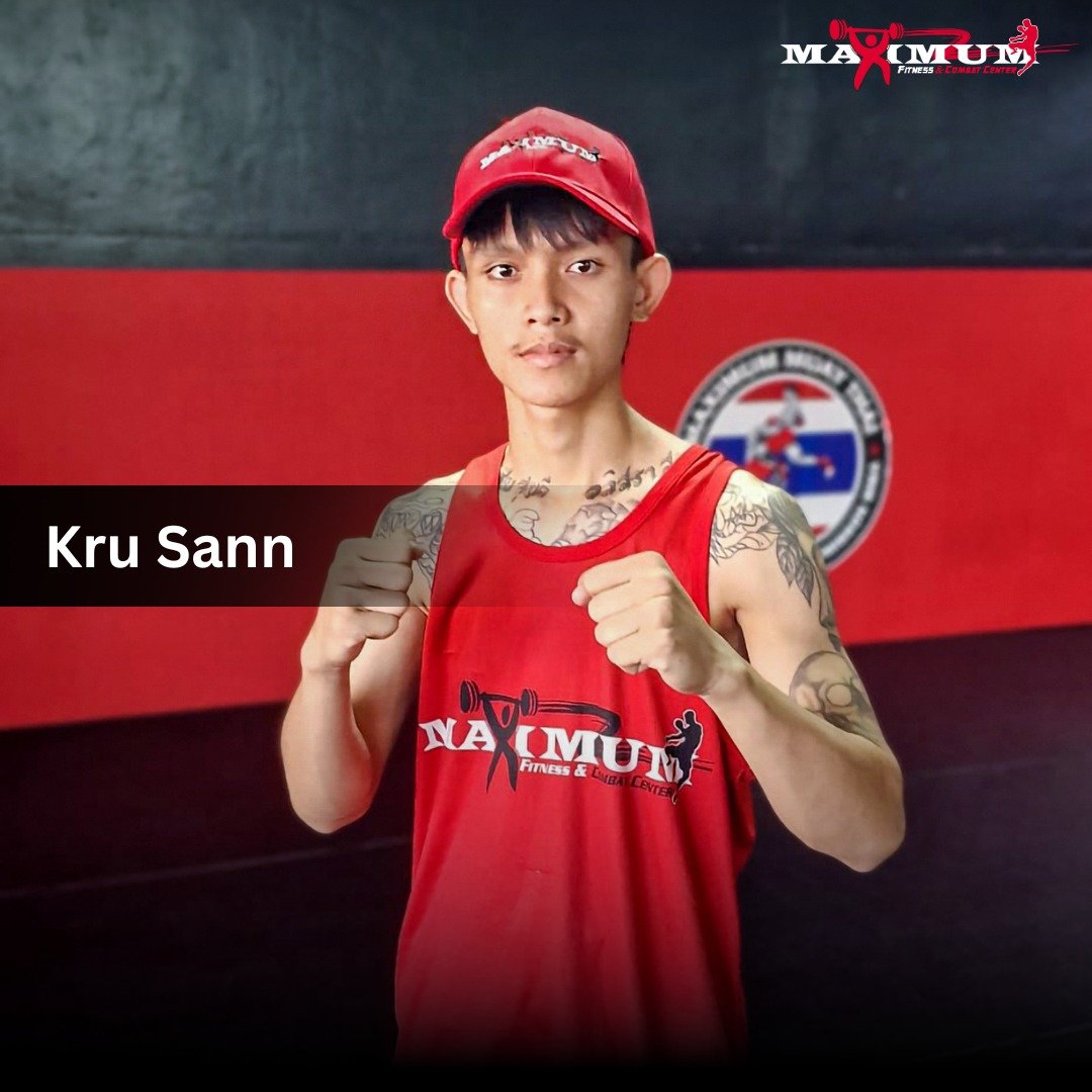 Kru Sann wearing a red tank top and cap with a martial arts or fitness gym logo, standing in a gym with a red and black background.