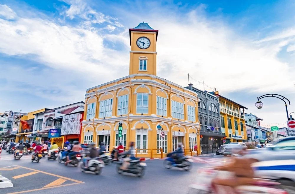 A yellow two-story building with a clock tower on top, situated on a busy street corner with many moving scooters and cars under a partly cloudy sky.