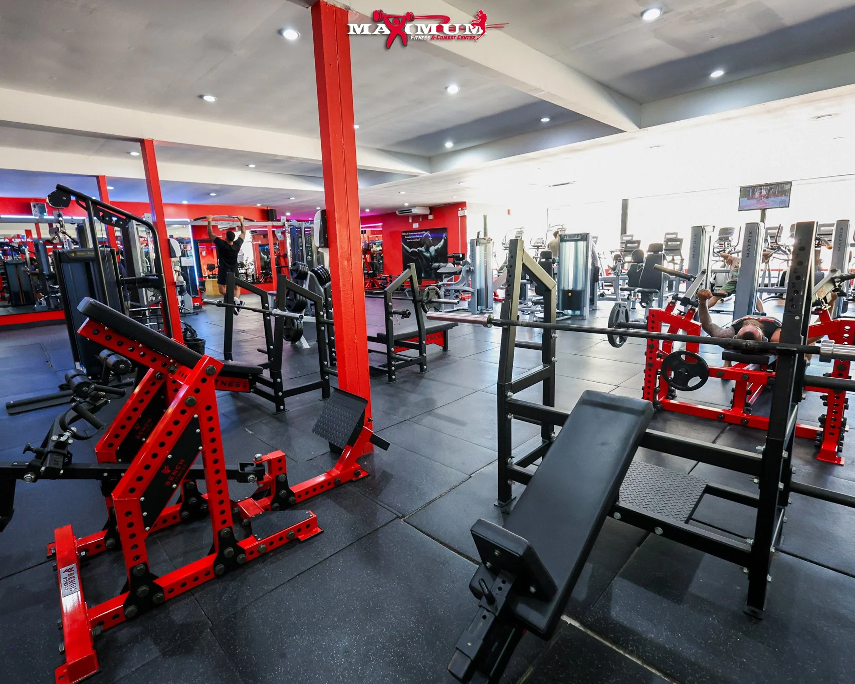 Interior of a fitness gym with various weightlifting and exercise equipment, including benches, barbells, and machines, with people working out in the background. The gym has a modern design with red and black accents.