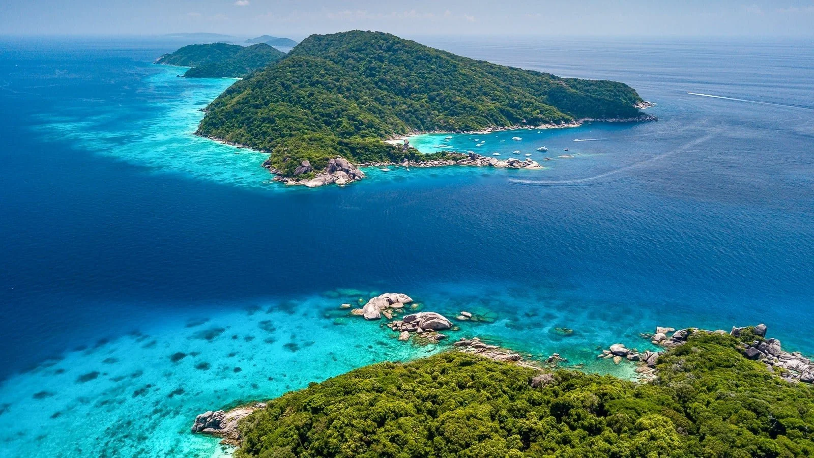 Aerial view of a tropical island with lush green trees, surrounded by bright blue ocean and coral reefs, with boats anchored near the shoreline.