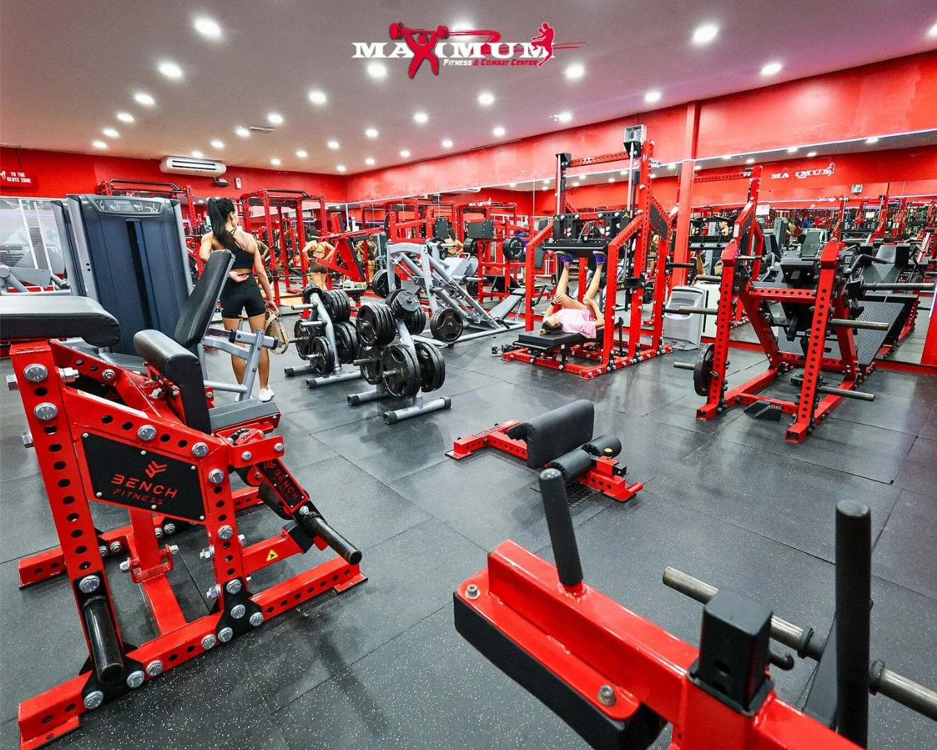 Interior of a gym with red and black workout equipment, weightlifting stations, and a mirrored wall. Two women are exercising; one lifting weights and the other lying on a bench. The gym has bright overhead lighting and a logo at the top center readi