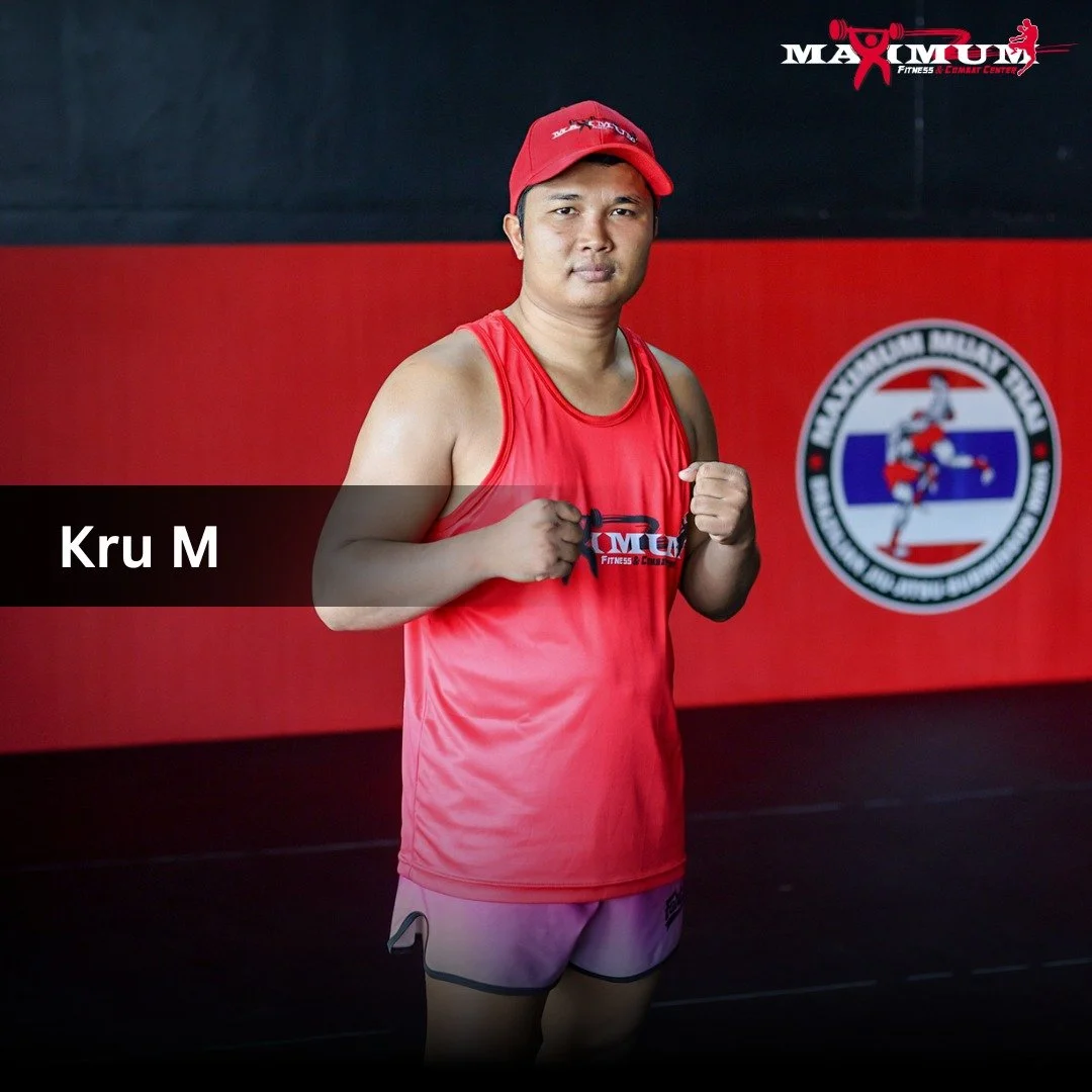 A man in a red sleeveless sports shirt and red cap standing in a boxing gym with one fist raised, posing confidently. The background features a black and red wall with a logo of Muay Thai boxing.