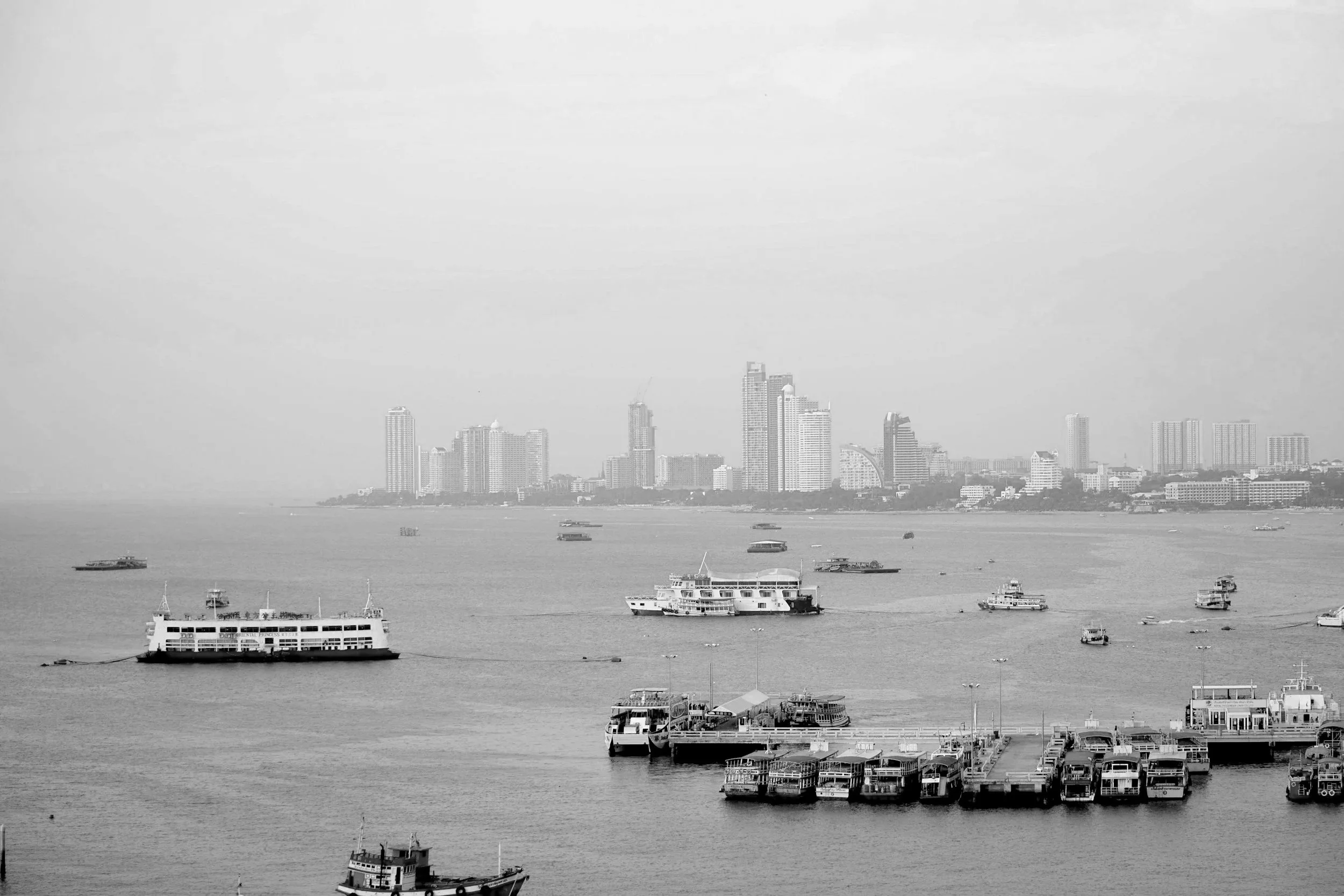 A black and white photo of a city skyline with high-rise buildings along a harbor, with boats and ferries in the water and a dock in the foreground.