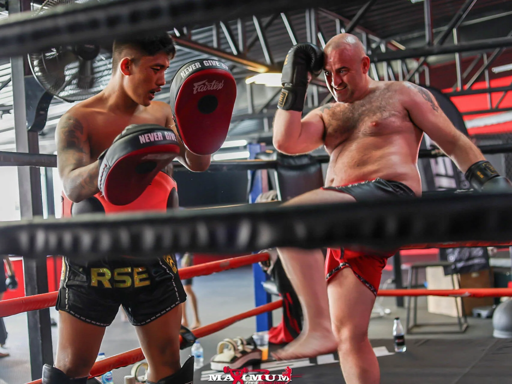 Two shirtless men in a boxing gym practicing martial arts. One is holding focus mitts, and the other is executing a kick.