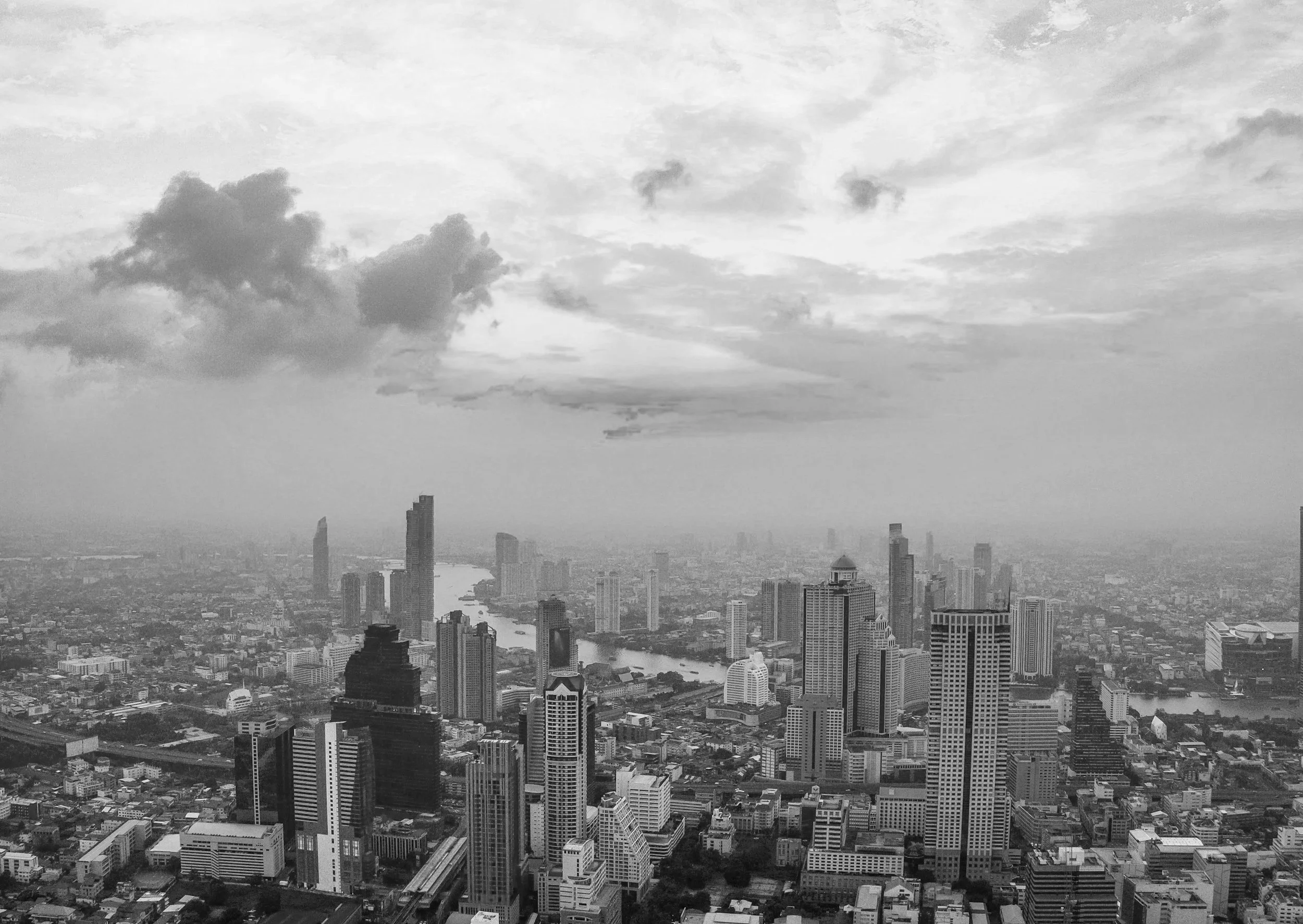 A black and white aerial view of a city skyline with tall skyscrapers, a river running through the city, and a cloudy sky.