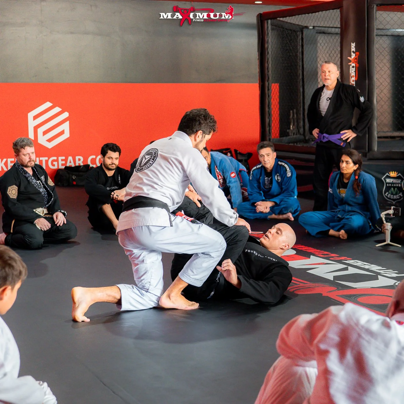 Martial arts instructor demonstrating a ground fighting technique to a student in a Brazilian Jiu-Jitsu class, with students and instructor watching attentively in a martial arts gym.