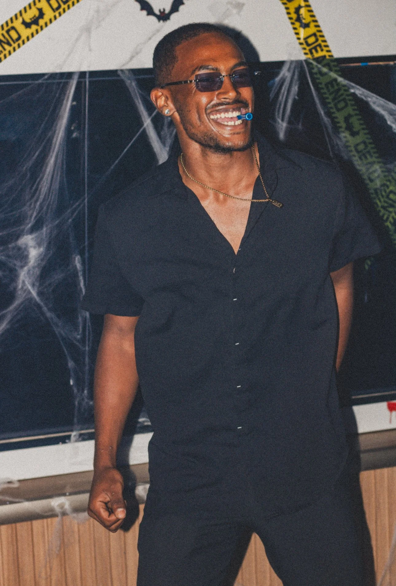 A man in black clothing, wearing sunglasses and a headset, smiling at a Halloween-themed party with spider web decorations and 'Caution' tape.