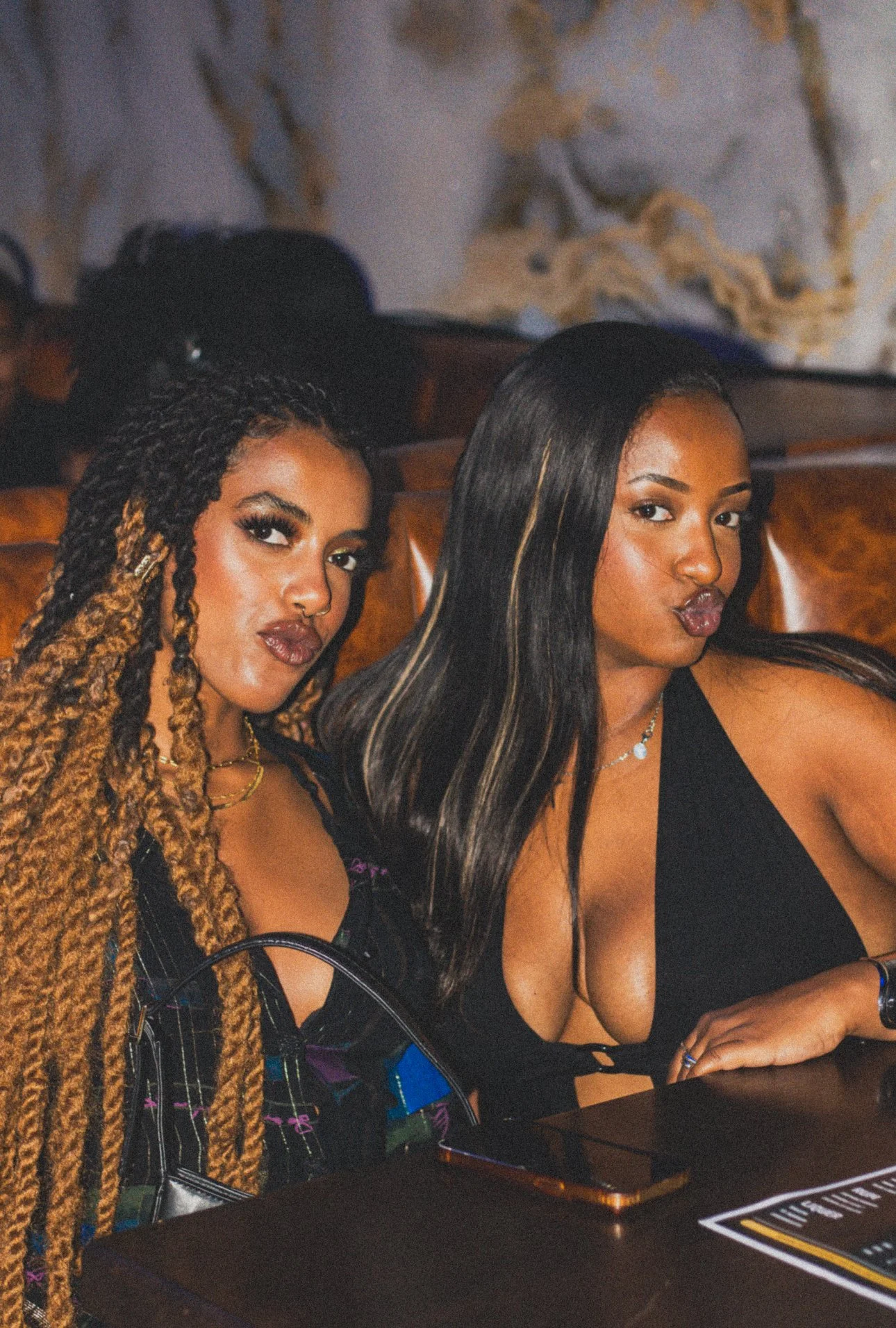 Two women sitting at a table, one with long braids and the other with straight black hair, both making pout faces for the camera.