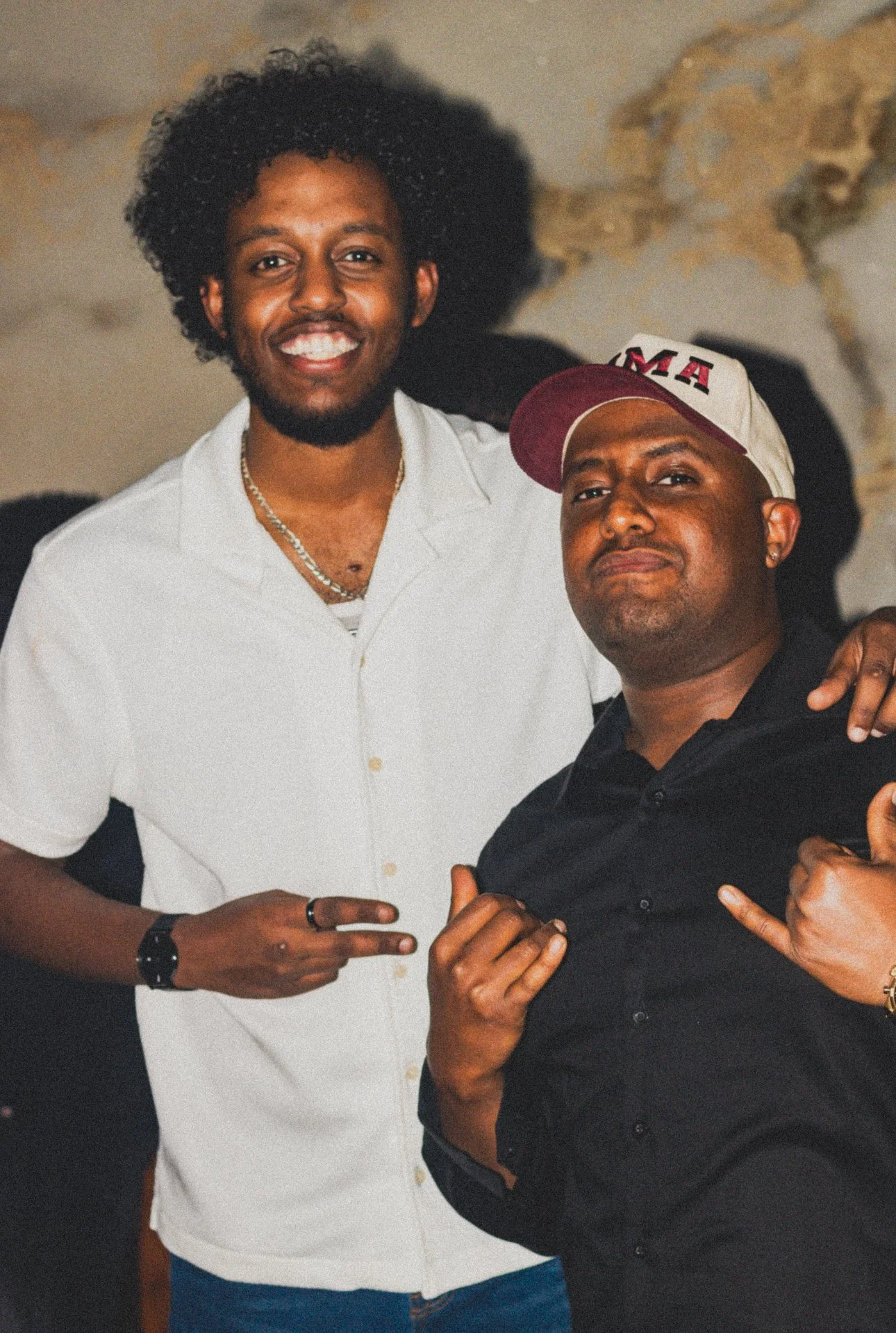 Two men posing together indoors with a stone wall in the background. The man on the left has curly hair, is smiling, and is pointing with his fingers. The man on the right is wearing a cap and making a hand gesture.