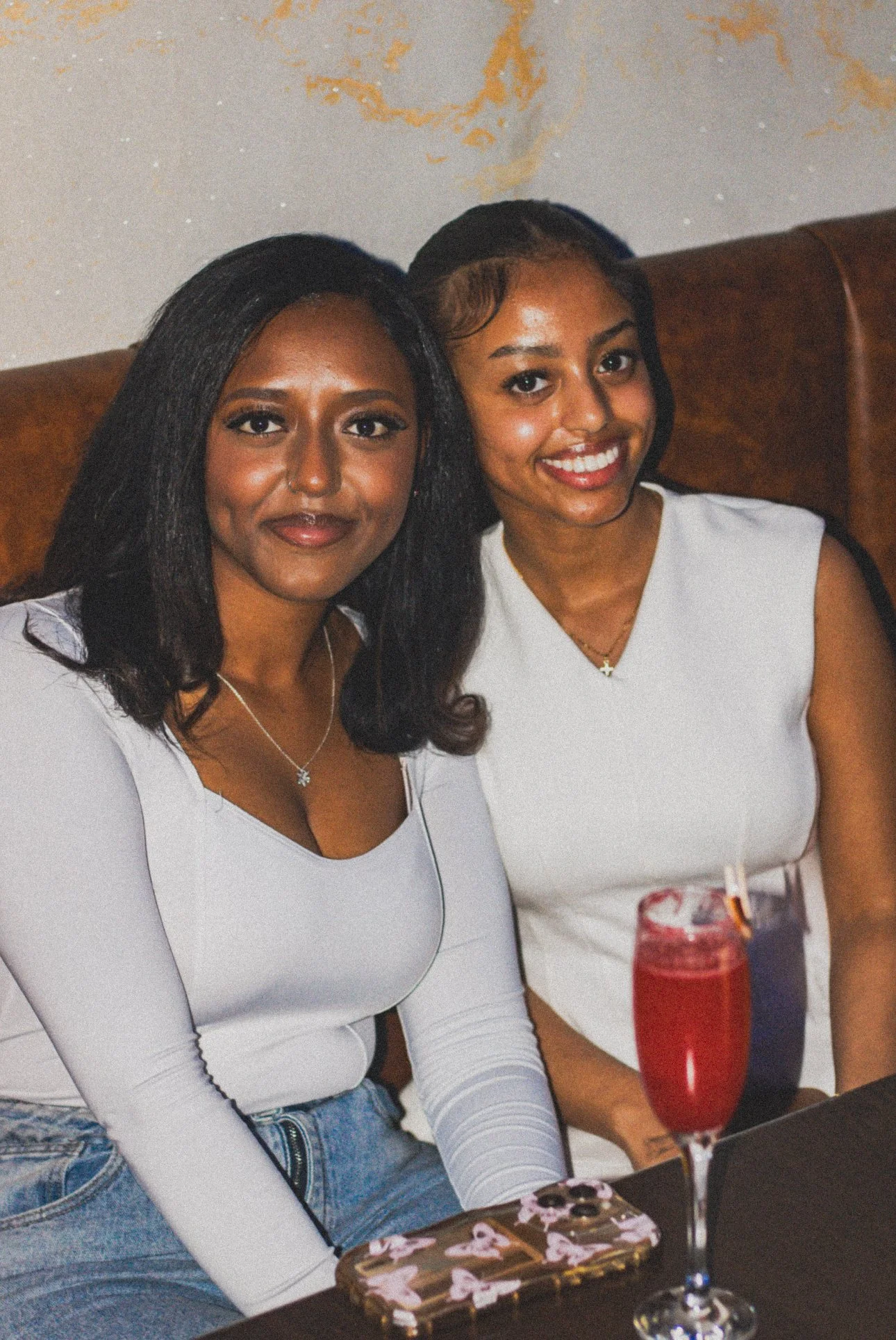 Two women sitting together at a dinner table, smiling at the camera, with a drink in front of one of them.