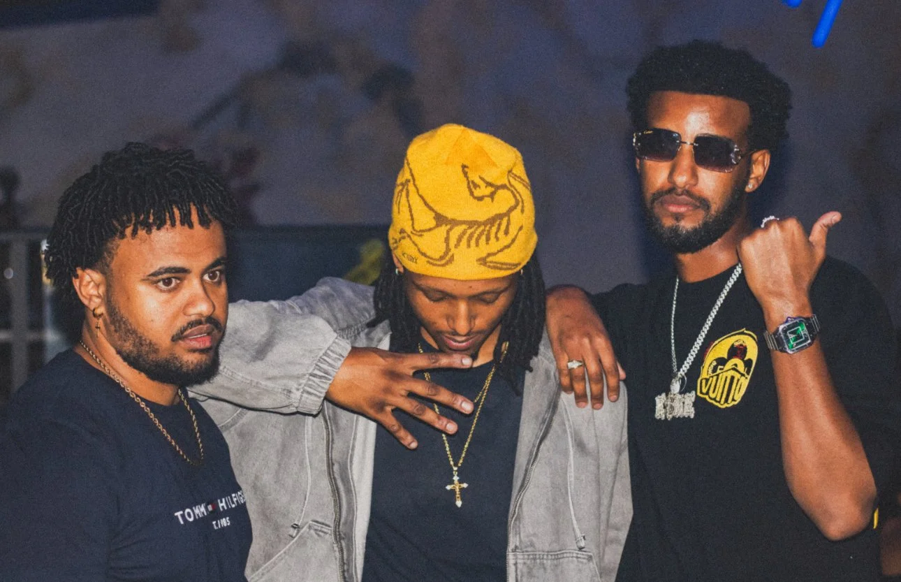 Three men standing close together with arms around each other, in a dimly lit setting. The man on the left has curly black hair and wears a black T-shirt and gold chain. The man in the middle wears a yellow headscarf and a gray jacket. The man on the