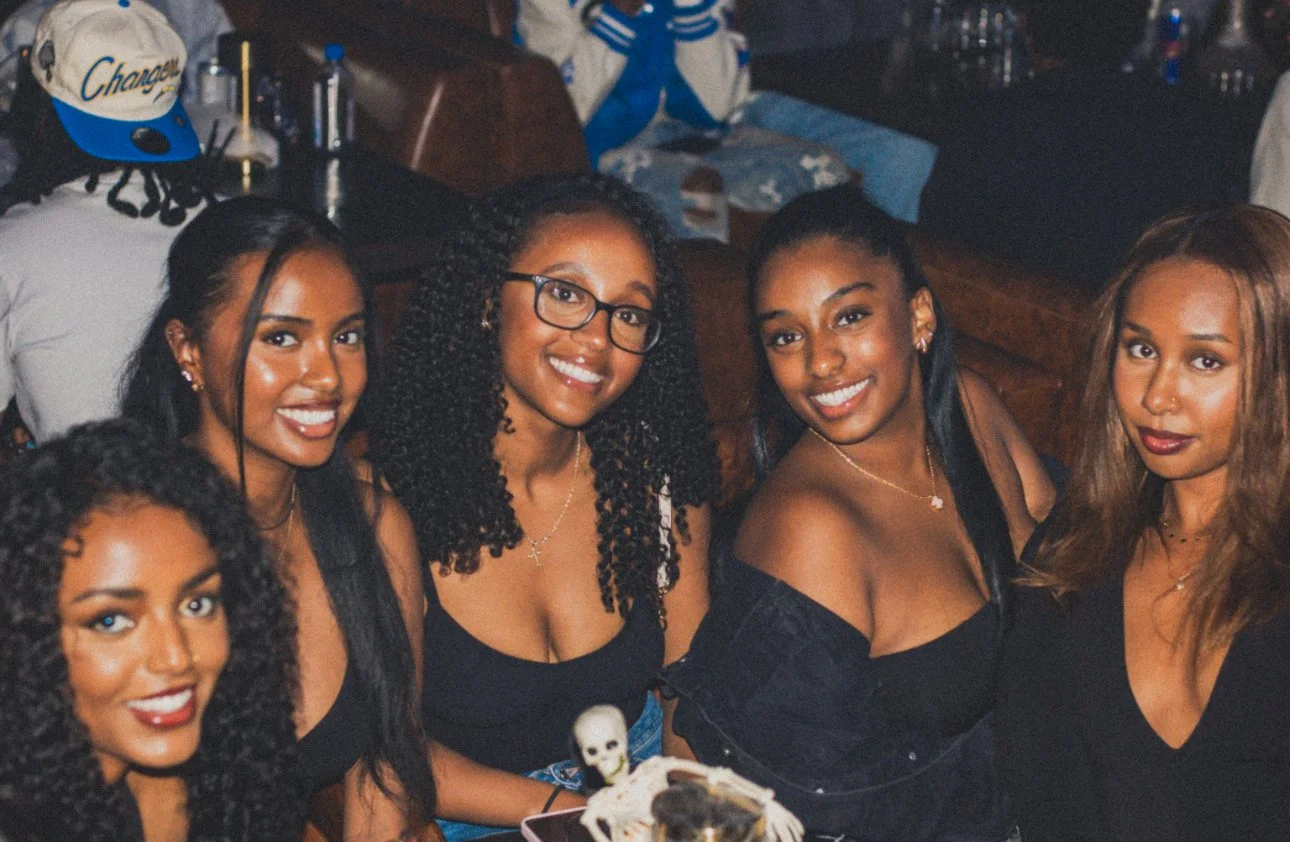 A group of five women with dark hair, dressed in black, gathered at a bar or restaurant, smiling at the camera, with a dessert topped with a skeleton decoration in front of them.