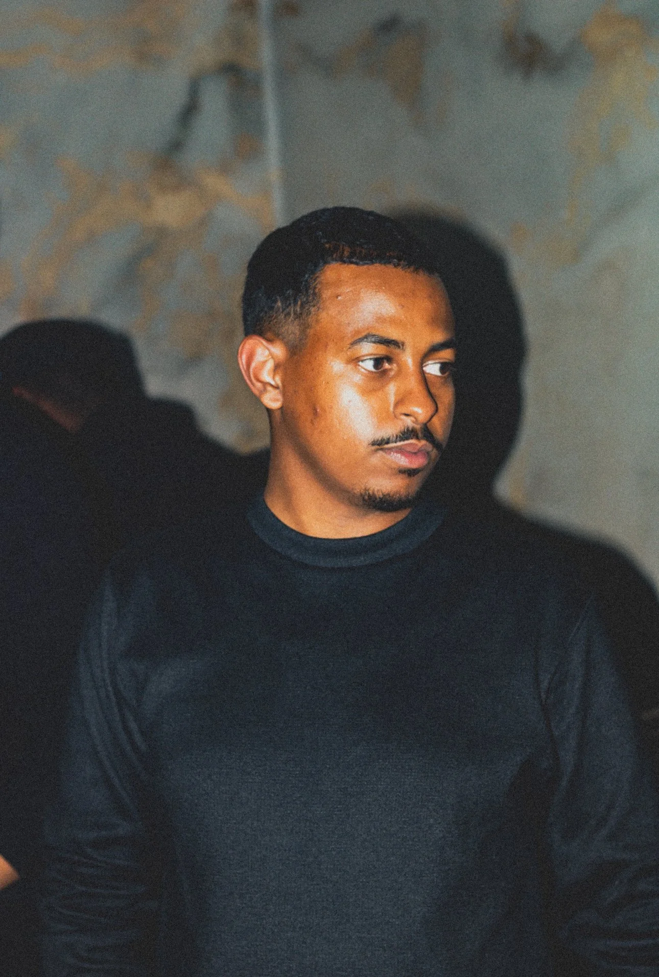 A young man with dark hair and a thin mustache, wearing a black shirt, looking to the side, with a textured wall in the background.