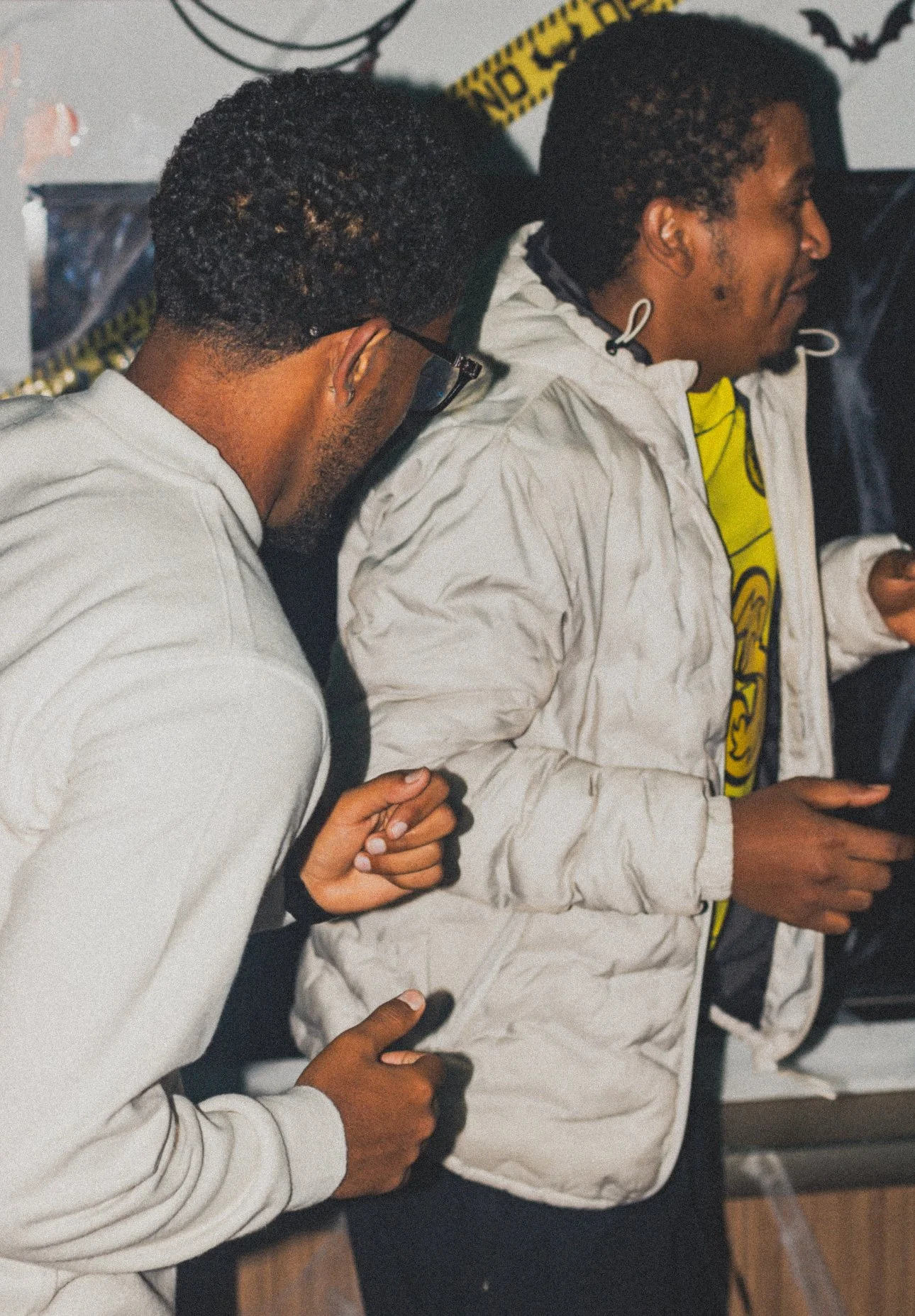Two young men, one with glasses and the other with curly hair, leaning in close together in a dimly lit room decorated for Halloween, with a warning tape and bat decoration visible in the background.