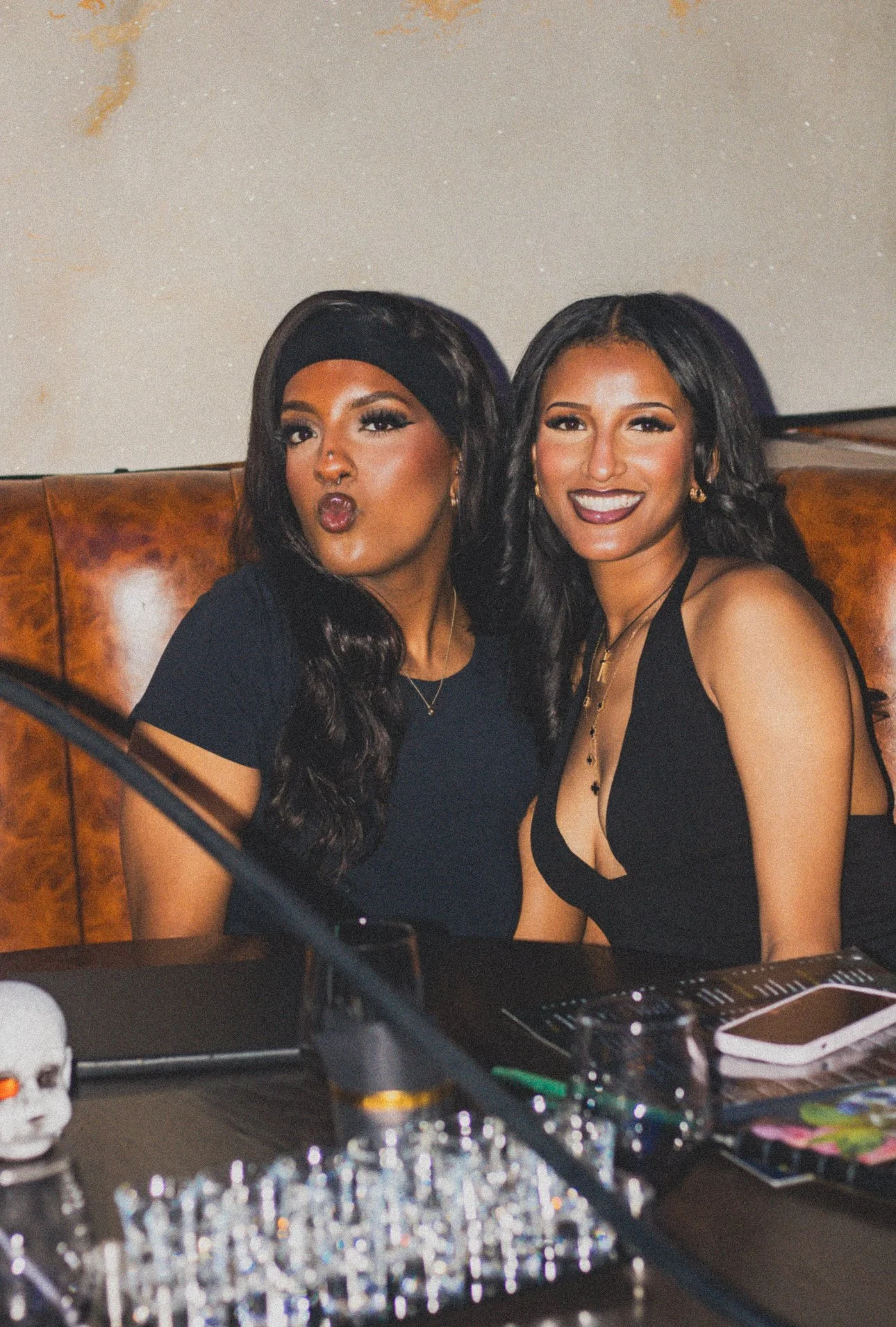 Two women sitting close together on a brown leather bench in a dimly lit setting, smiling and making a playful face. One woman has long dark hair and is wearing a black headband and dark clothing, while the other has long sparking hair and is dressed