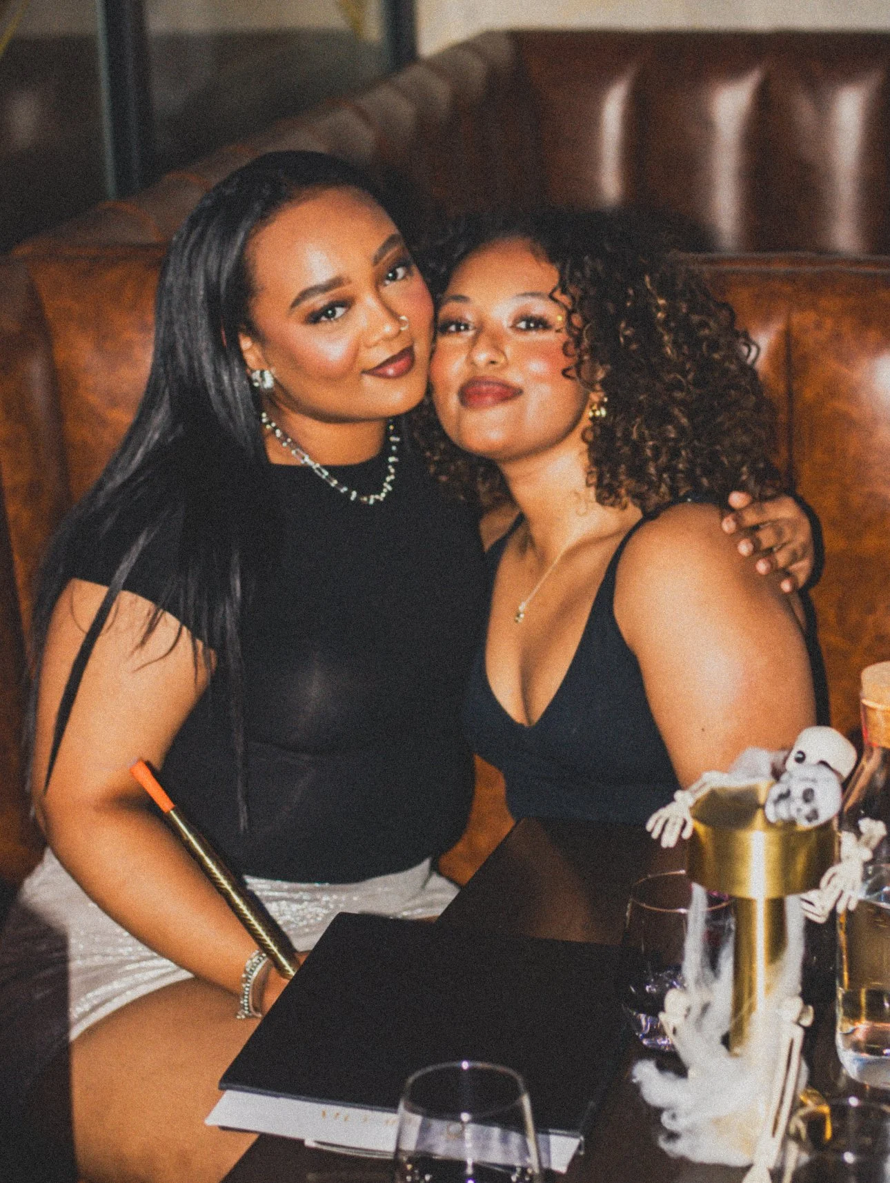 Two women sitting close together at a table, smiling and embracing. One has long black hair and the other curly brown hair, both dressed in black. The table has a black notebook, glasses, and decorations, including a golden drum with skeleton decorat