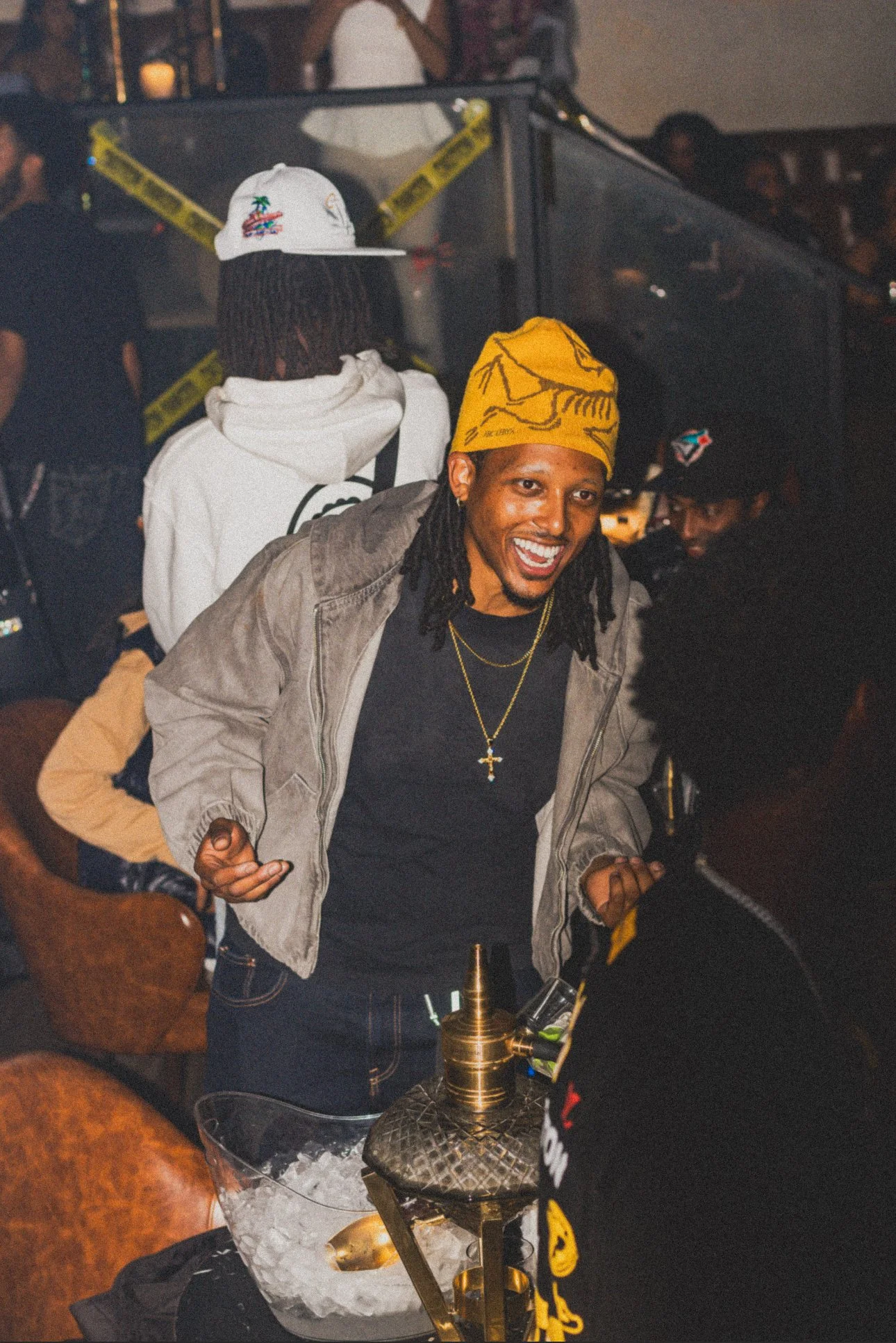 A man with dreadlocks wearing a yellow hat, black T-shirt, and gray jacket smiling and leaning forward at a party or social gathering.