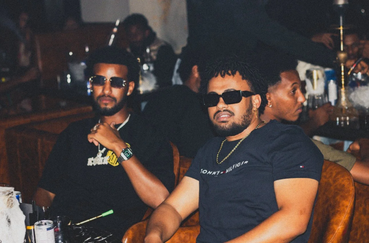 Two men wearing black shirts and sunglasses sitting at a table in a smoky bar or lounge, with other people in the background.
