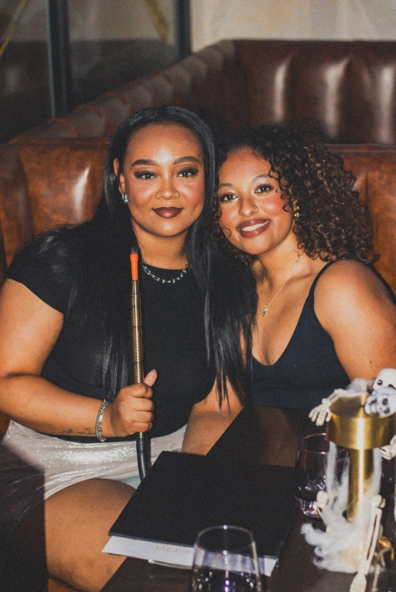 Two women sitting close together, smiling at the camera in a dimly lit setting, with one holding a hookah pipe.