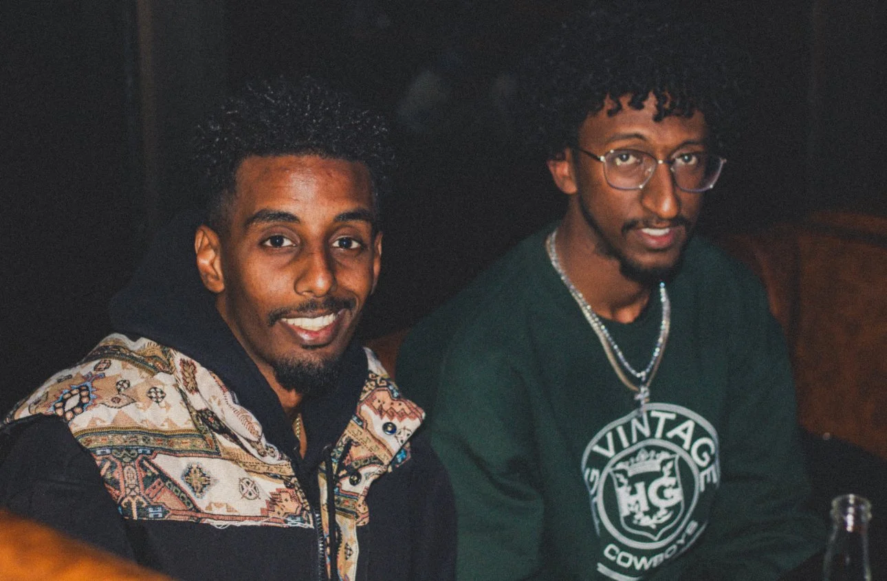 Two young men sitting together indoors, one smiling at the camera and wearing a patterned jacket, the other with glasses and wearing a black sweatshirt with a circular logo, in a dimly lit setting.
