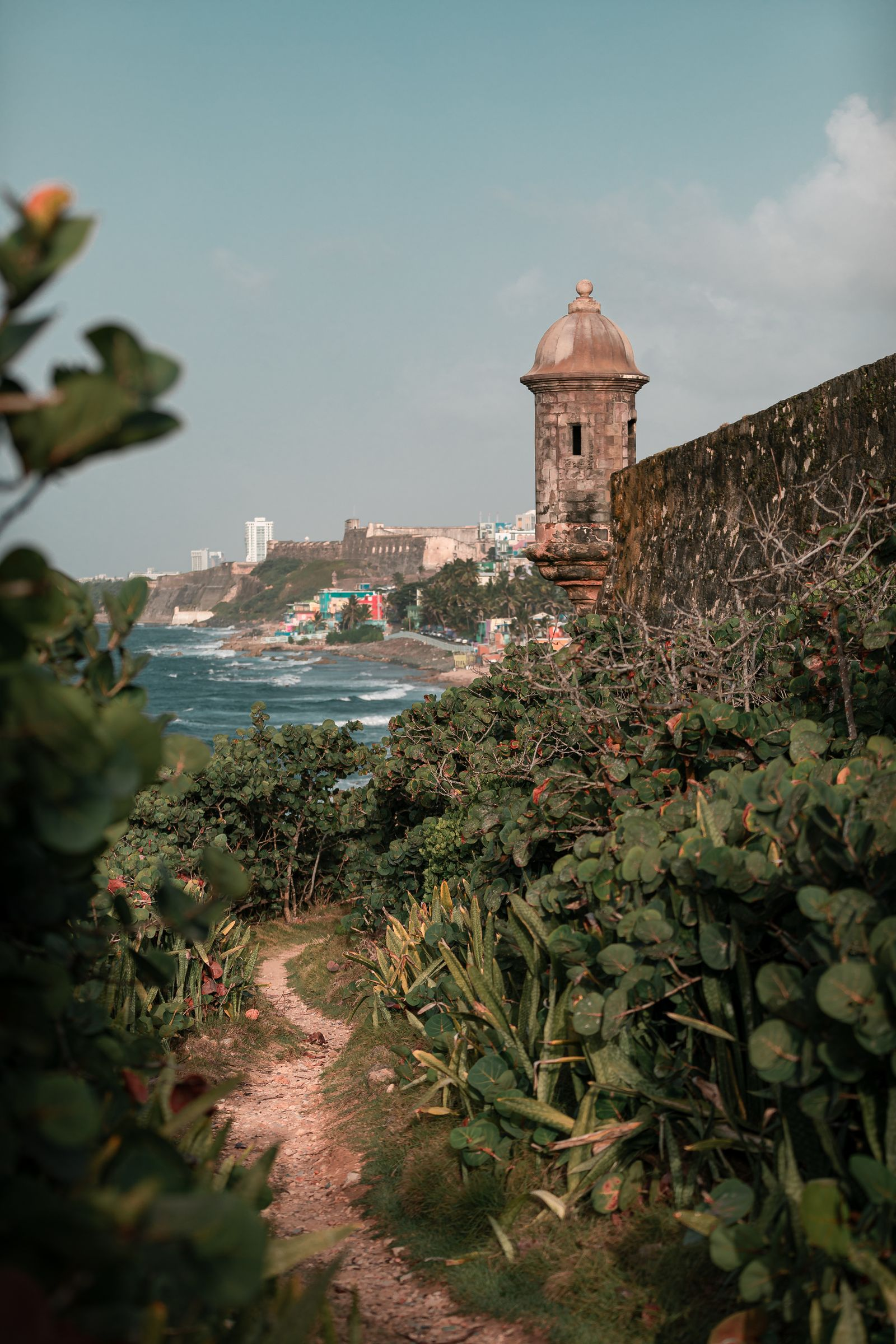A narrow dirt path lined with lush green bushes leads towards a stone wall and lookout tower on a coastal cliff. The ocean waves crash against the shore in the background, with colorful buildings and a historic fortress further along the coastline. The sky is partly cloudy.