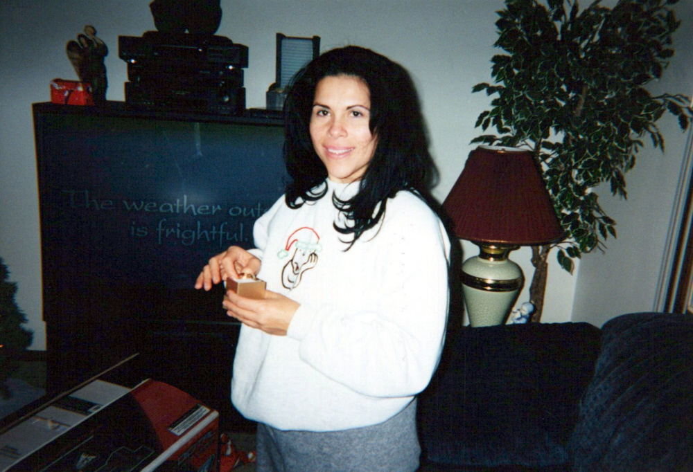 A woman with long black hair standing in a living room, holding a small box. There is a TV behind her with text on the screen that reads 'The weather out is frightful.'