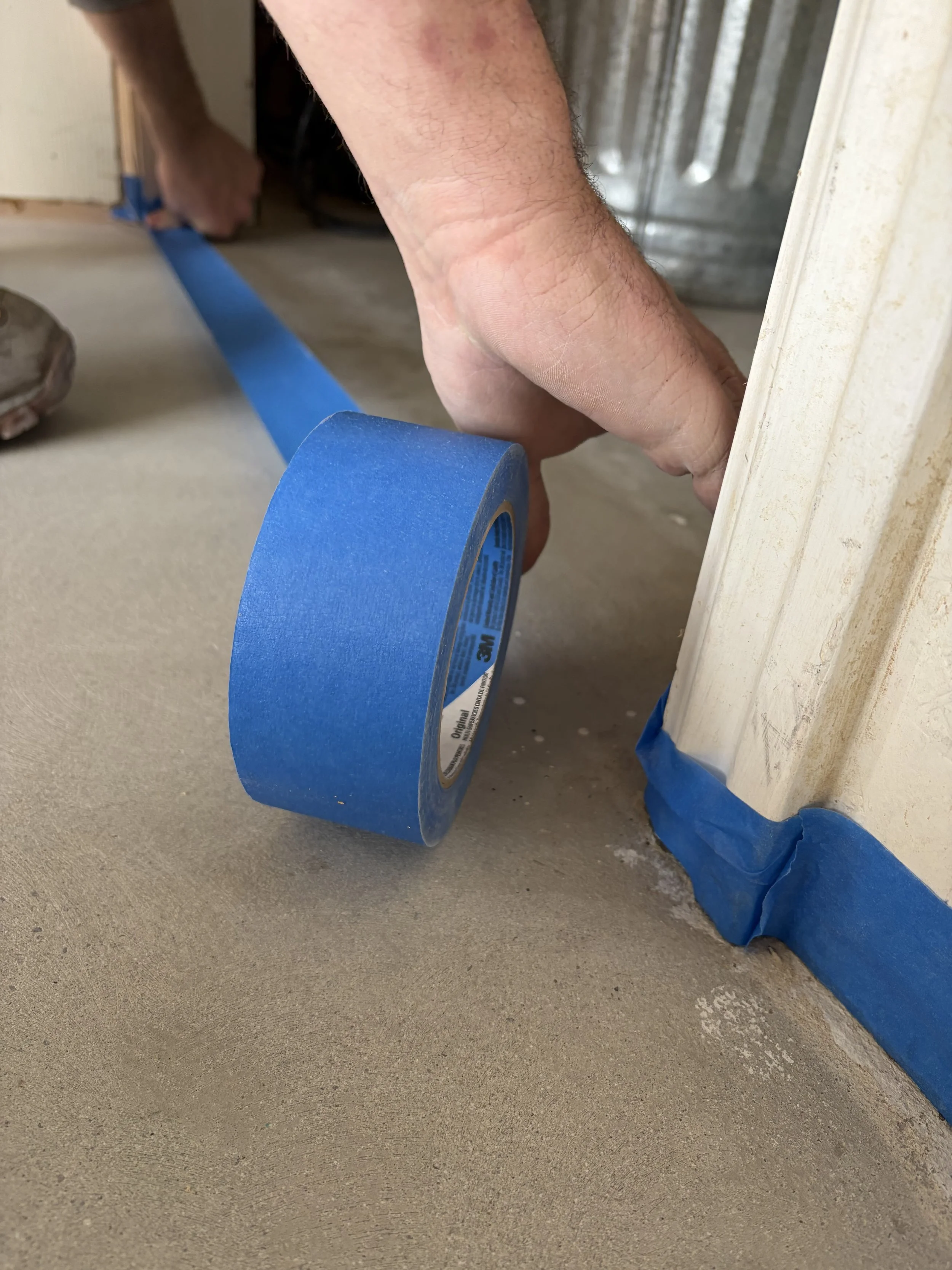 A person's hand applying blue painter's tape along the edge of a doorway to protect the wall while painting.