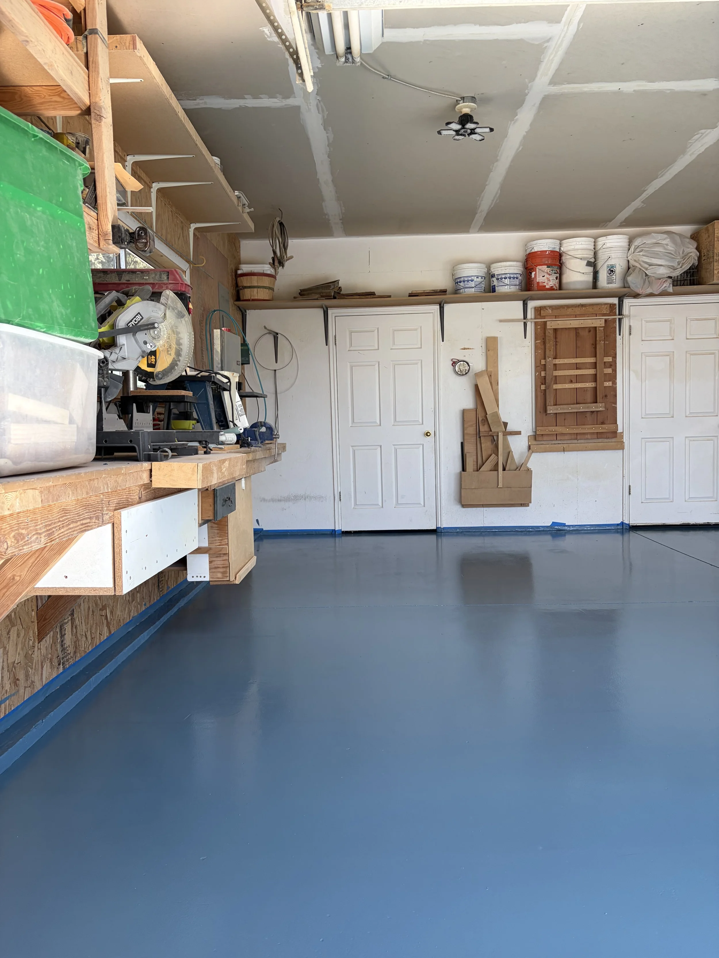 A clean garage with a freshly painted smooth blue floor, white garage doors, and a workbench area with tools and storage on the left, and shelves with buckets and supplies on top of the garage doors on the back wall.