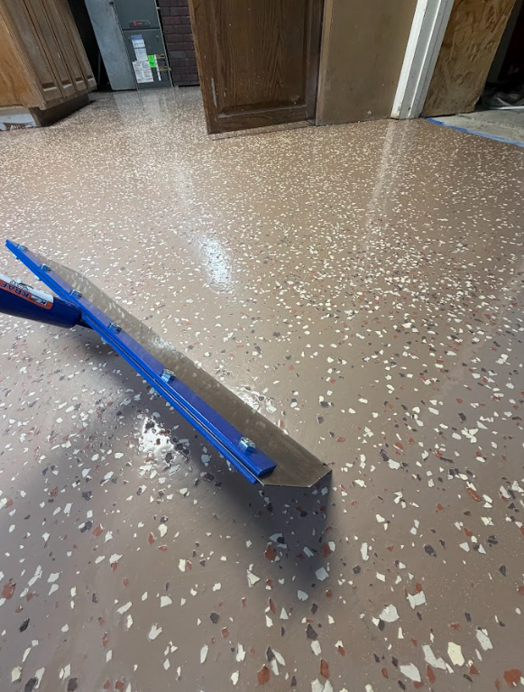 A floor sander with a long blue handle resting on a speckled brown and white terrazzo floor in a room with wood cabinets and a door.