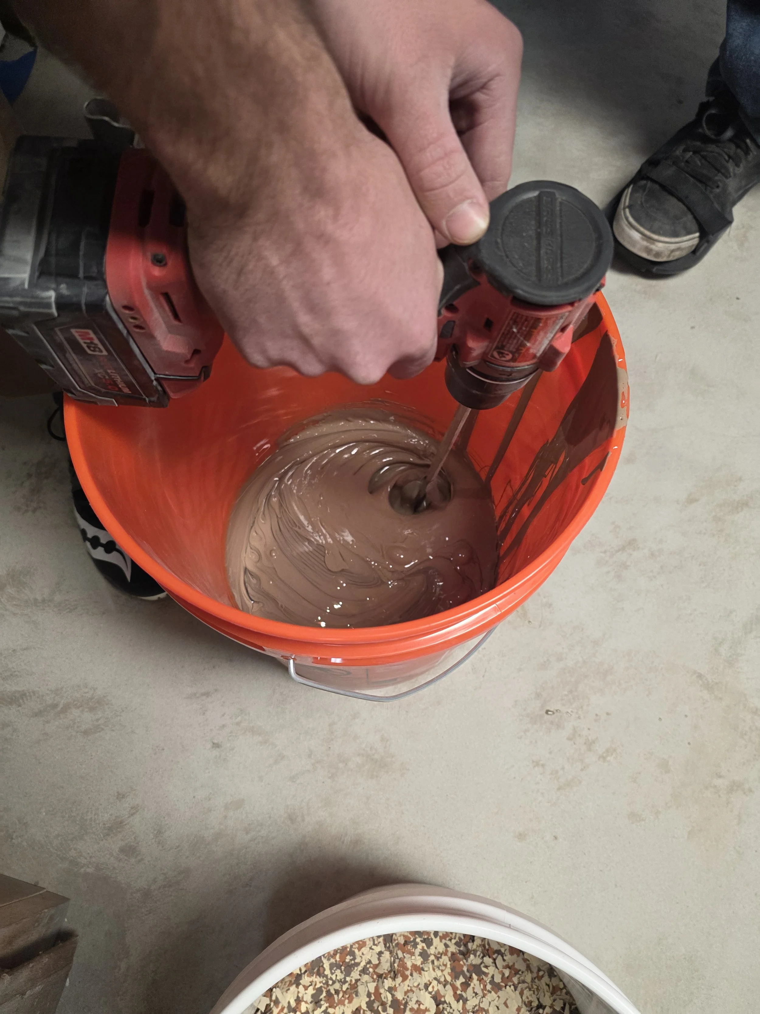 A person using a red cordless power drill to mix chocolate-colored substance in an orange bucket. A person’s hand and part of their shoe are visible, with a concrete floor underneath.