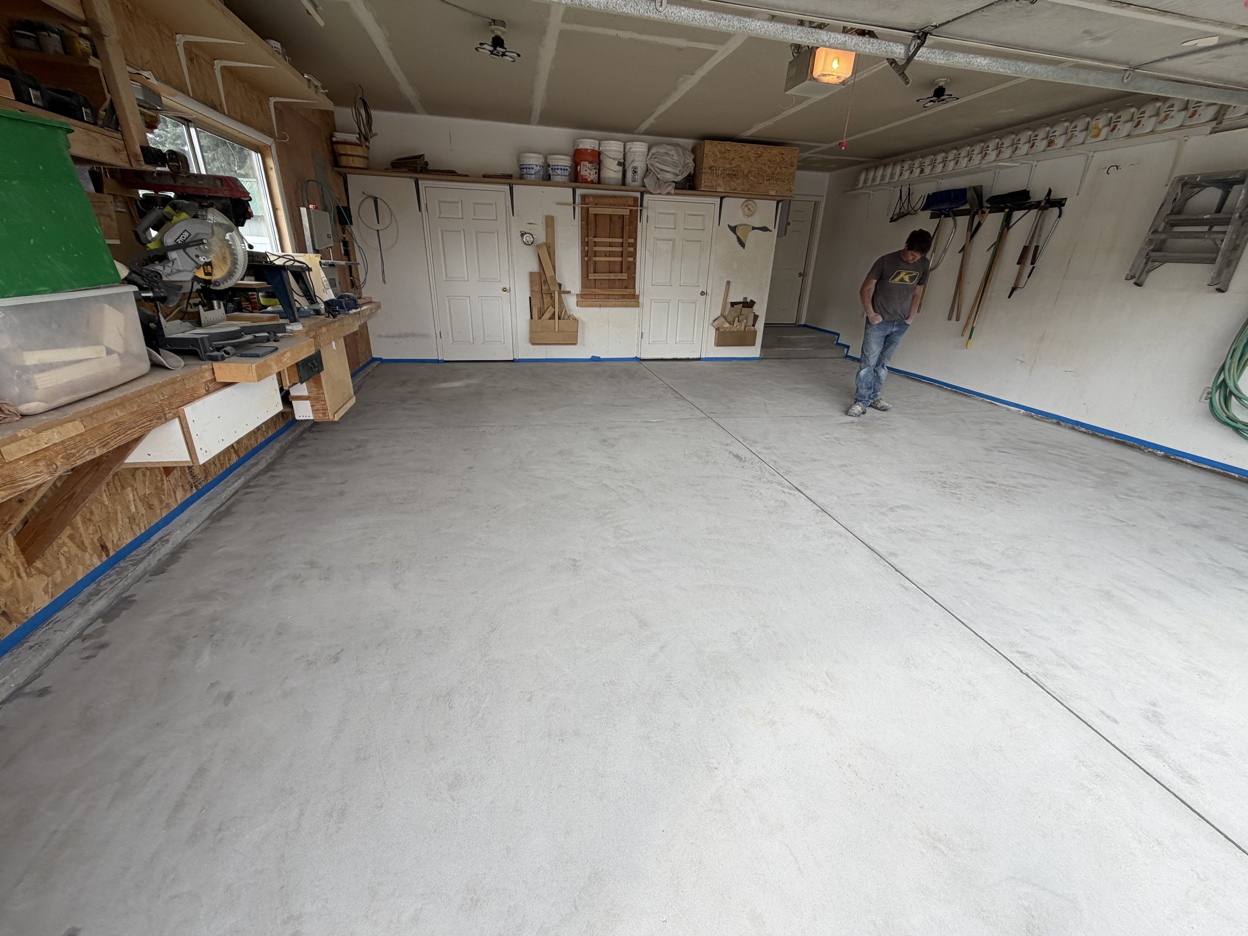 A spacious garage with a concrete floor in development, with various tools and equipment along the walls, a person standing near the right wall, and organized storage on shelves and hooks.