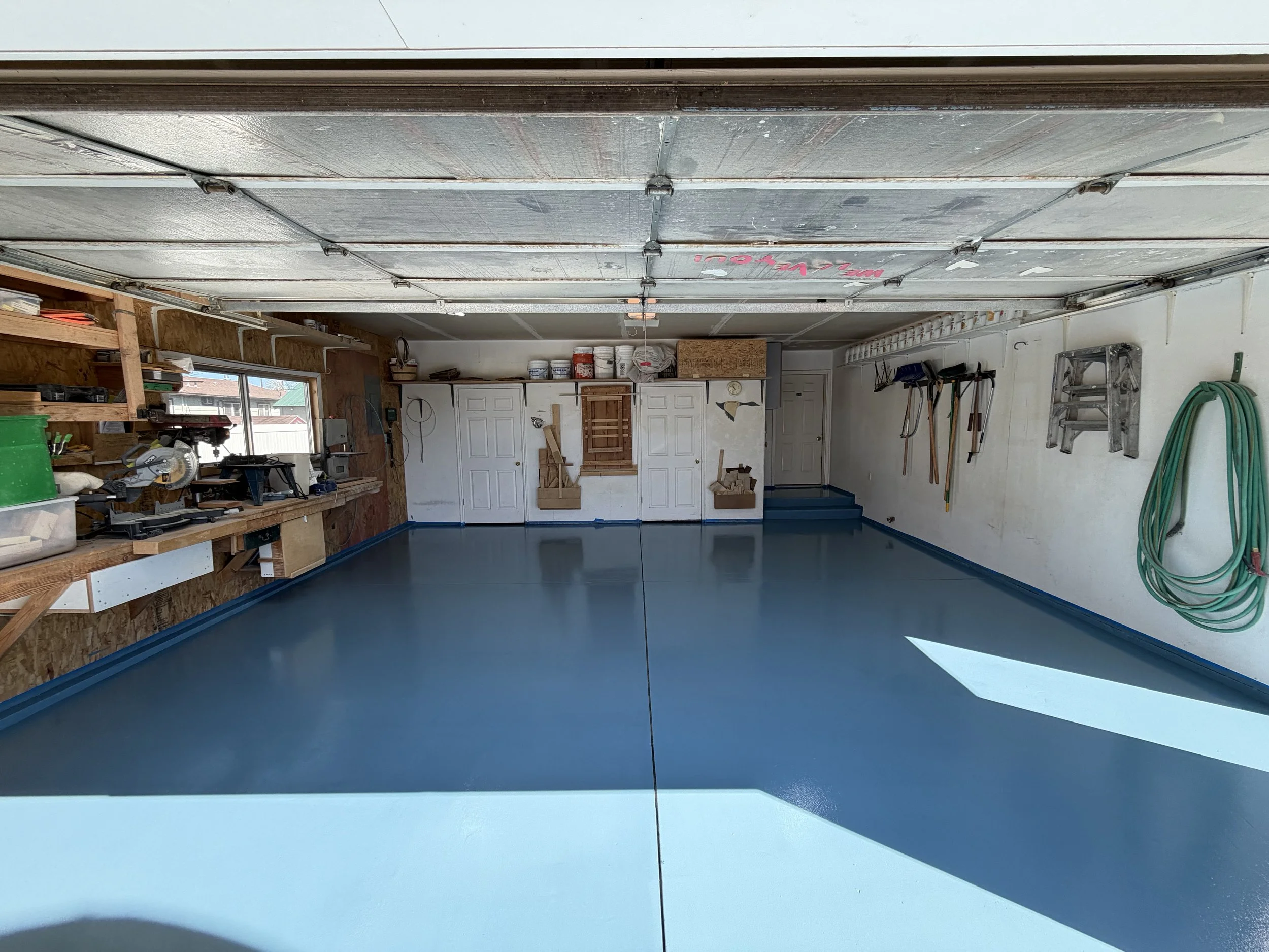 Empty garage with blue epoxy floor, tools organized on wall-mounted racks, windows on the left, cabinets, and storage on the back wall with sunlight streaming in.