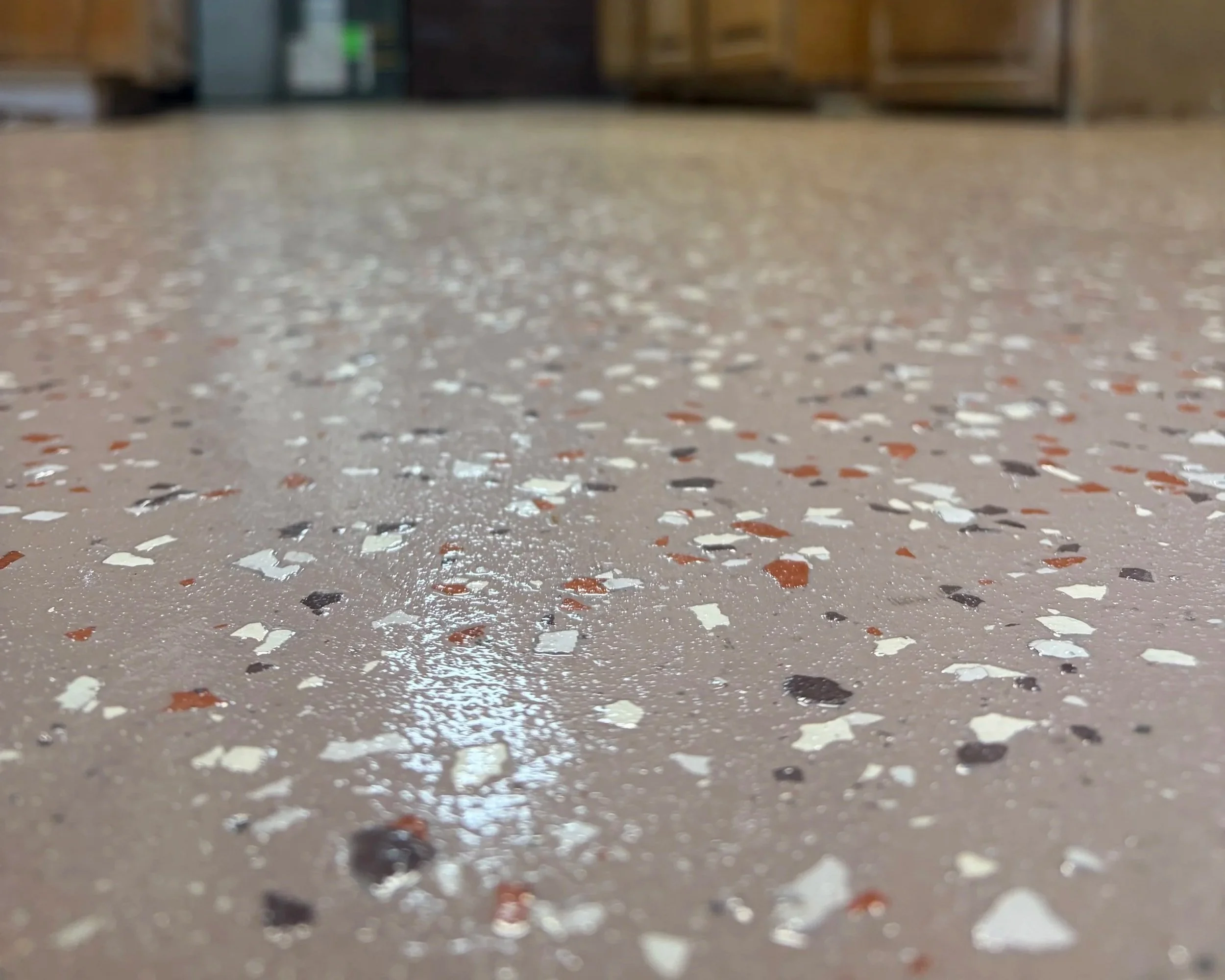 Close-up of a speckled terrazzo floor with various colored chips, reflecting light, with blurred background of wood and metal furniture.