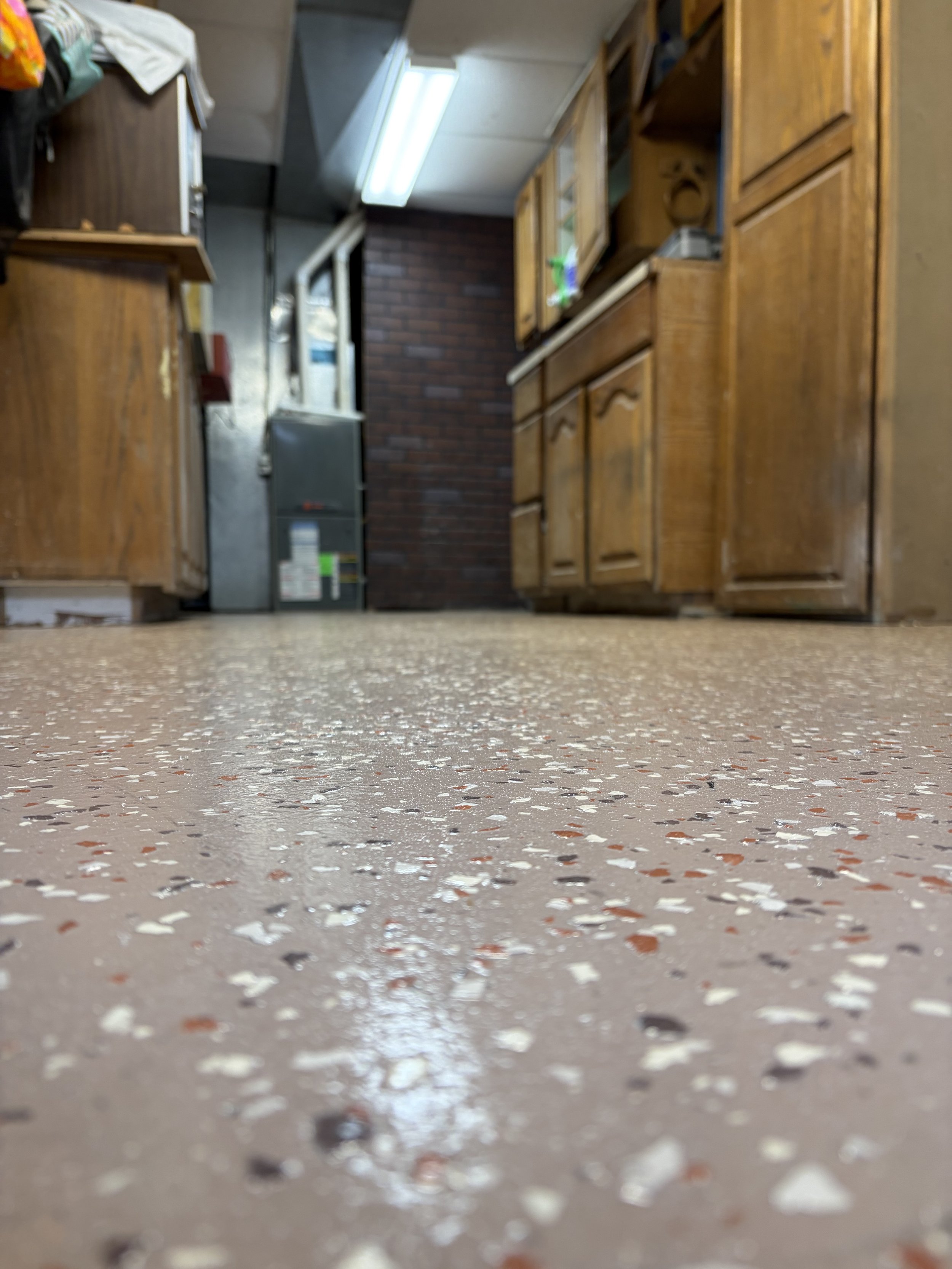 Close-up of a speckled beige and brown floor in a kitchen with wooden cabinets and a brick wall in the background.