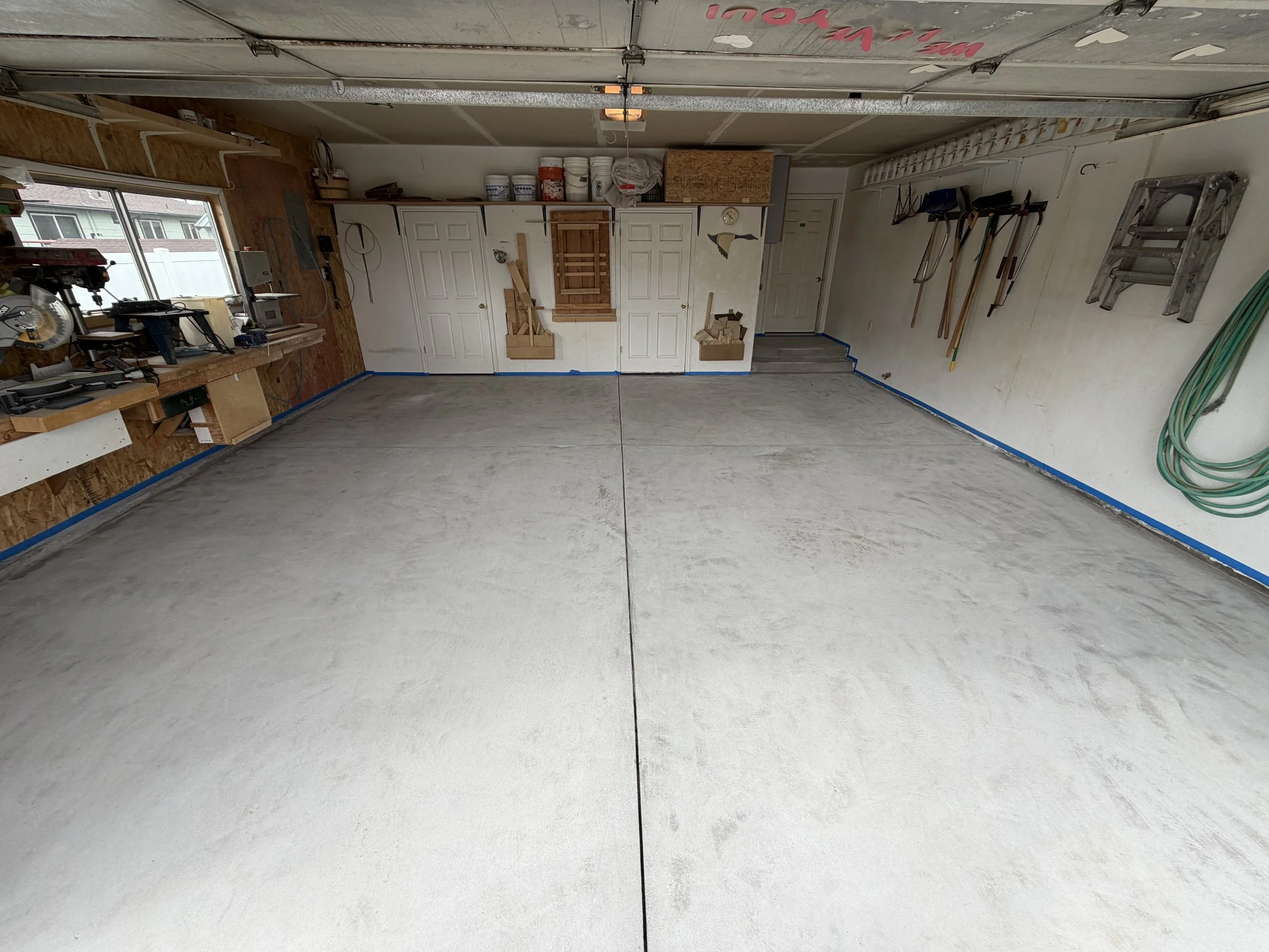 Empty garage with concrete floor and tools hanging on the wall, window on the left, and storage cabinets at the back.