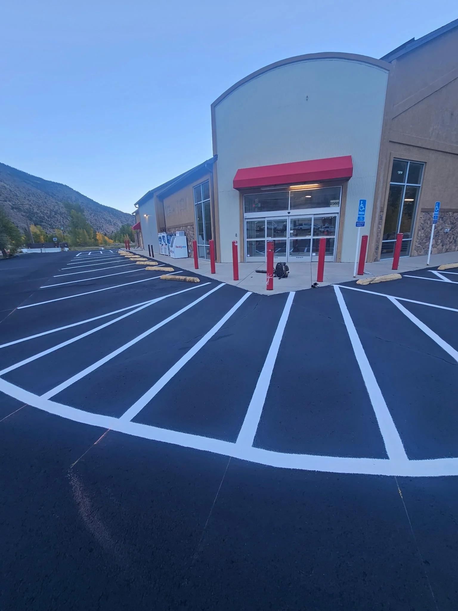 An empty parking lot in front of a retail store with multiple handicapped parking spaces and red posts near the entrance, surrounded by mountains and a clear blue sky.