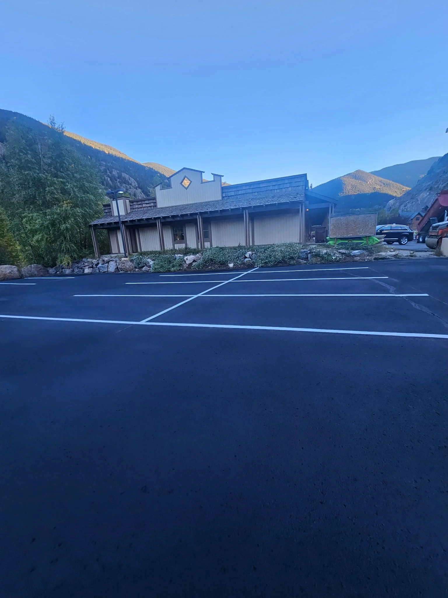 Empty parking lot with white marked lines, wooden building with decorative architectural features, surrounded by mountains and trees, under a clear blue sky.