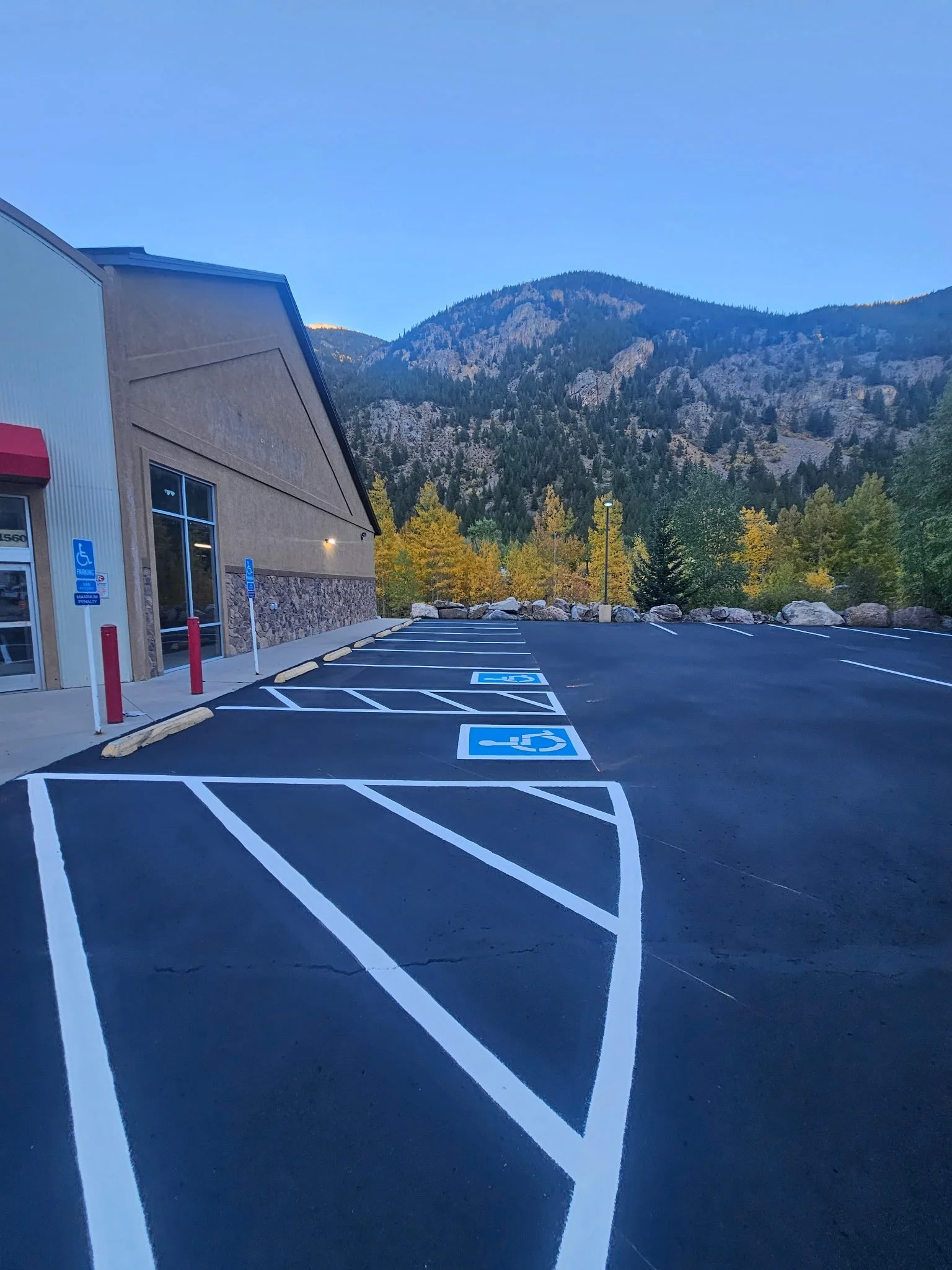 Empty parking lot with designated handicapped parking spaces marked with blue and white symbols, in front of a building with a mountain landscape and trees in the background.