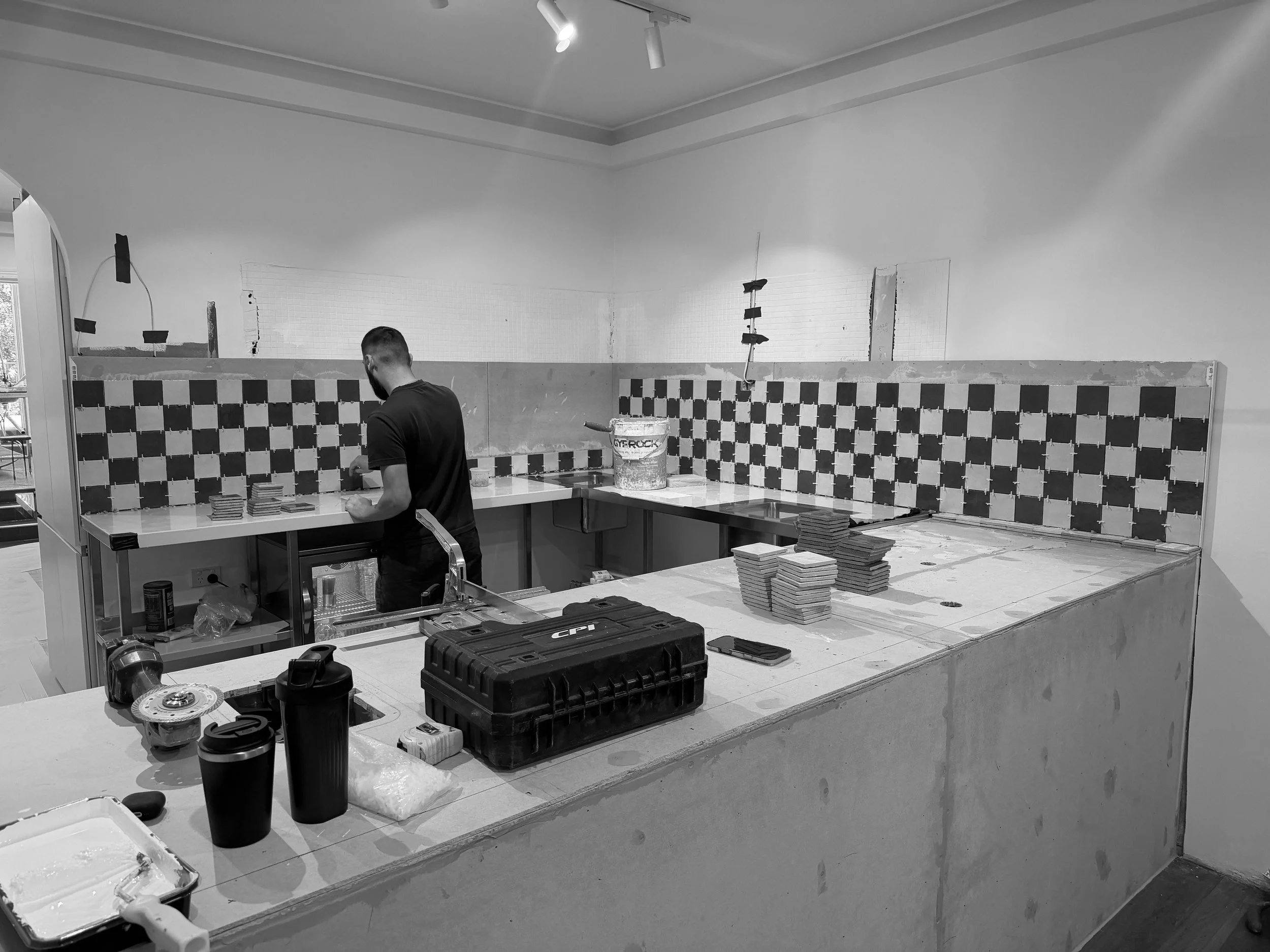 A man is working on a construction or renovation project in a room. The room has a checkered tile pattern on the lower part of the wall. Construction tools, materials, and supplies are scattered on the counter and floor.