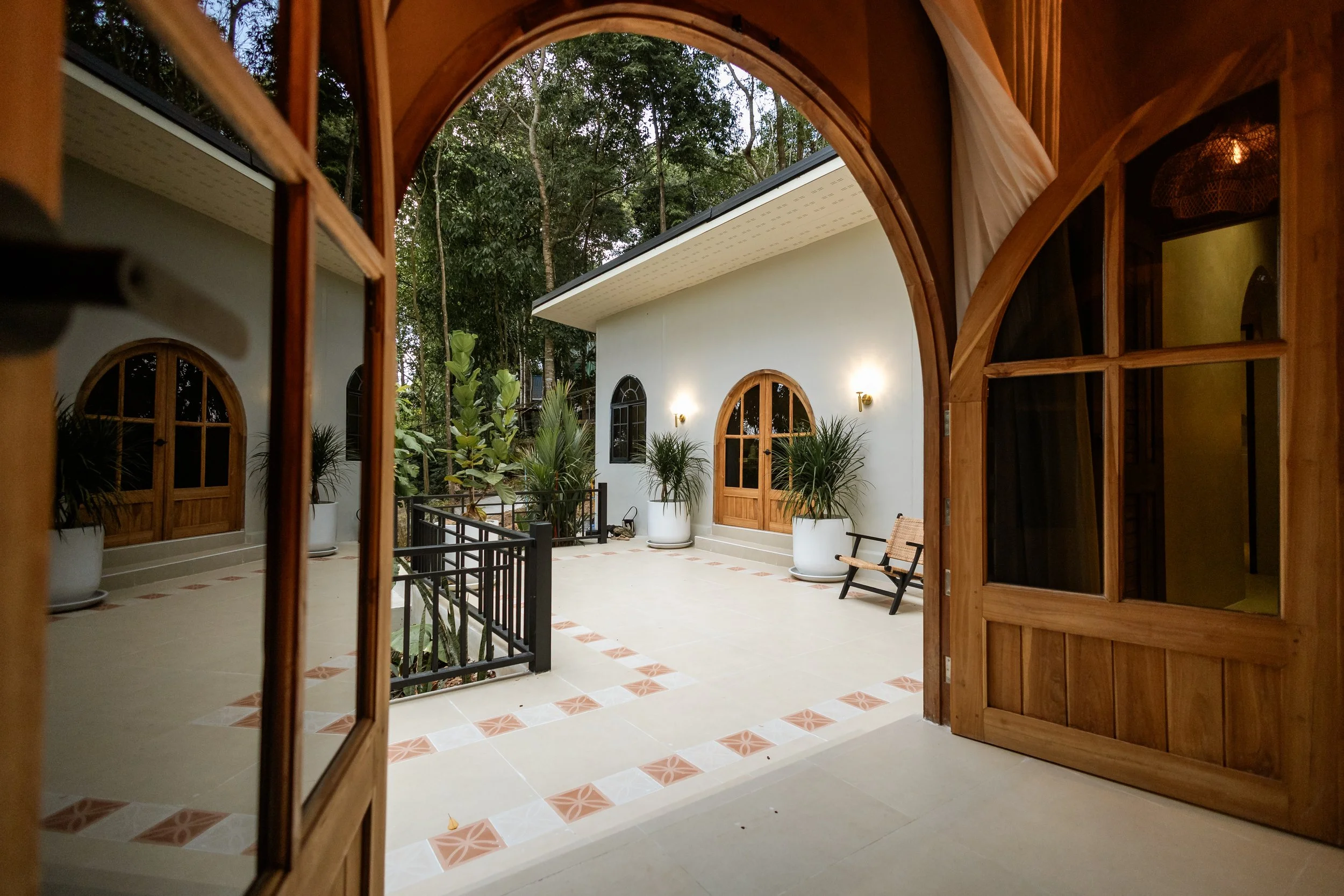 View of a sunny outdoor patio with white walls, arched windows, potted plants, and a wooden bench, seen through a wooden door with glass panels.